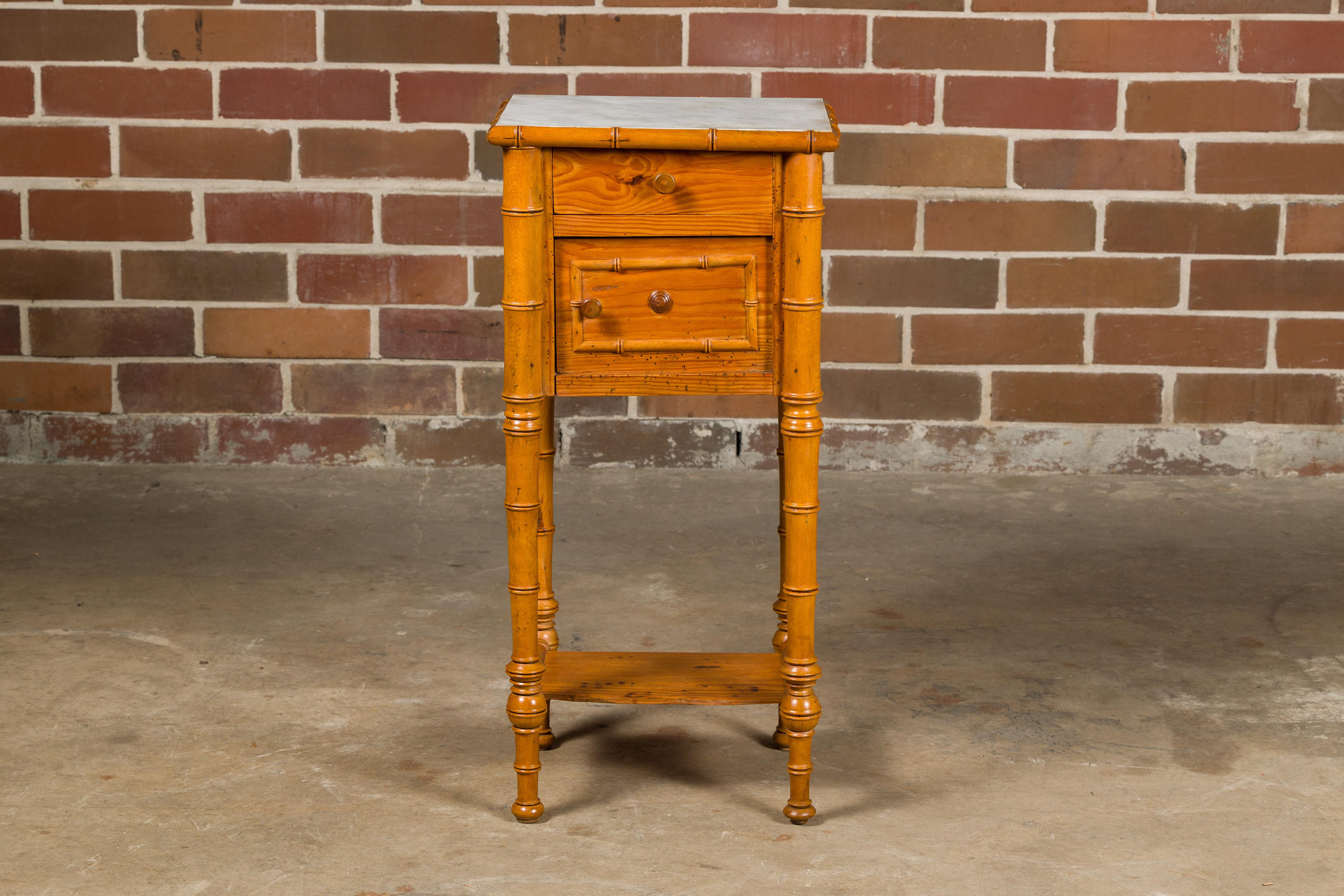 A French faux bamboo pine side table from the 19th century with white marble top, single drawer as well as door and lower shelf. Immerse yourself in the quaint charm of this 19th-century French faux bamboo pine side table, a piece that beautifully