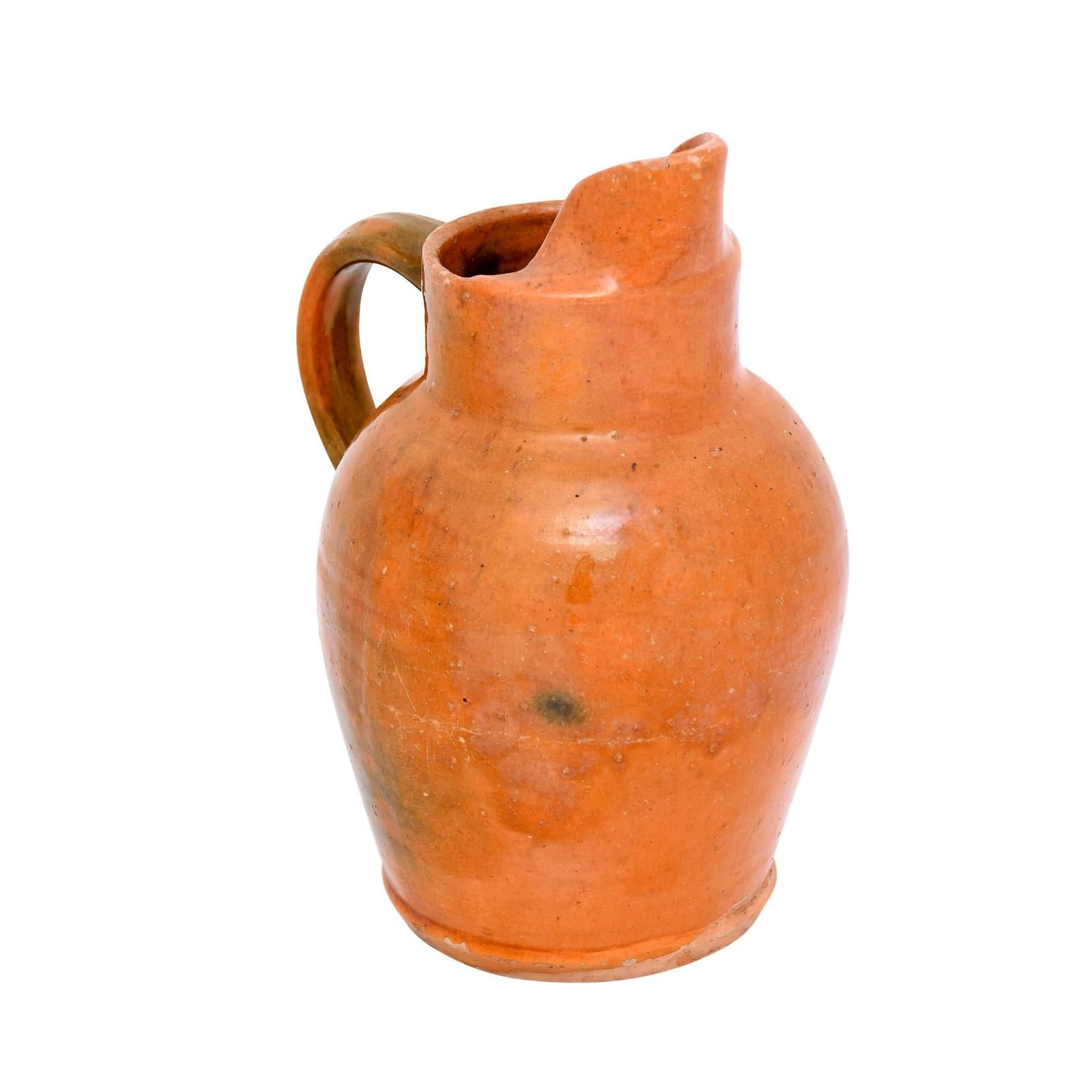 A French orange glazed pottery jug from the 19th century, made to store and serve wine or apple cider, with back handle and front spout. Step back in time with this French 19th-century pottery jug that embodies the rustic charm of traditional French