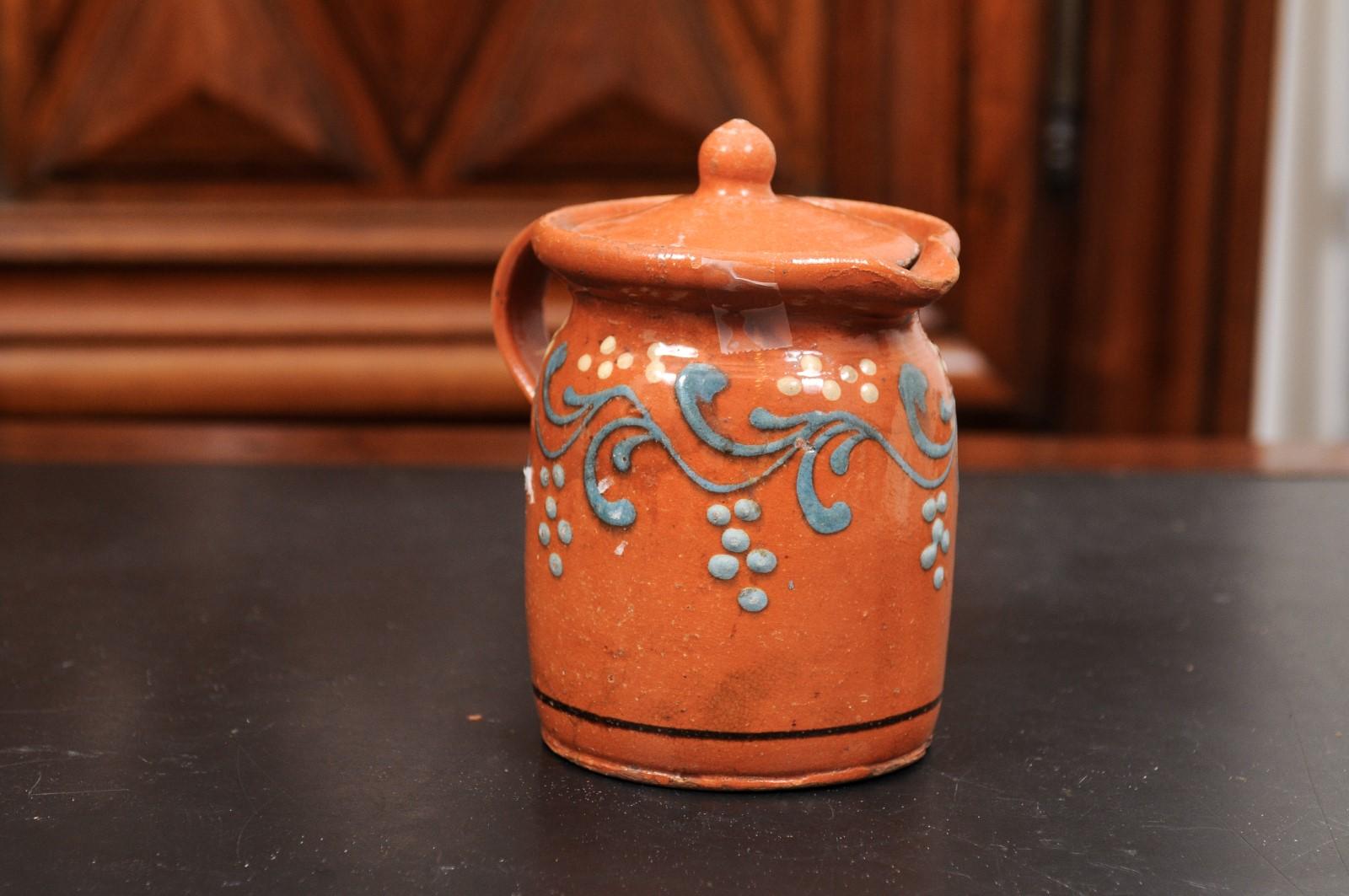 A French pottery lidded pitcher from the 19th century, with orange, cream and blue glaze and foliage motifs. Created in France during the 19th century, this pottery pitcher charms us with its vivid colors and foliage motifs. Presenting a handle in