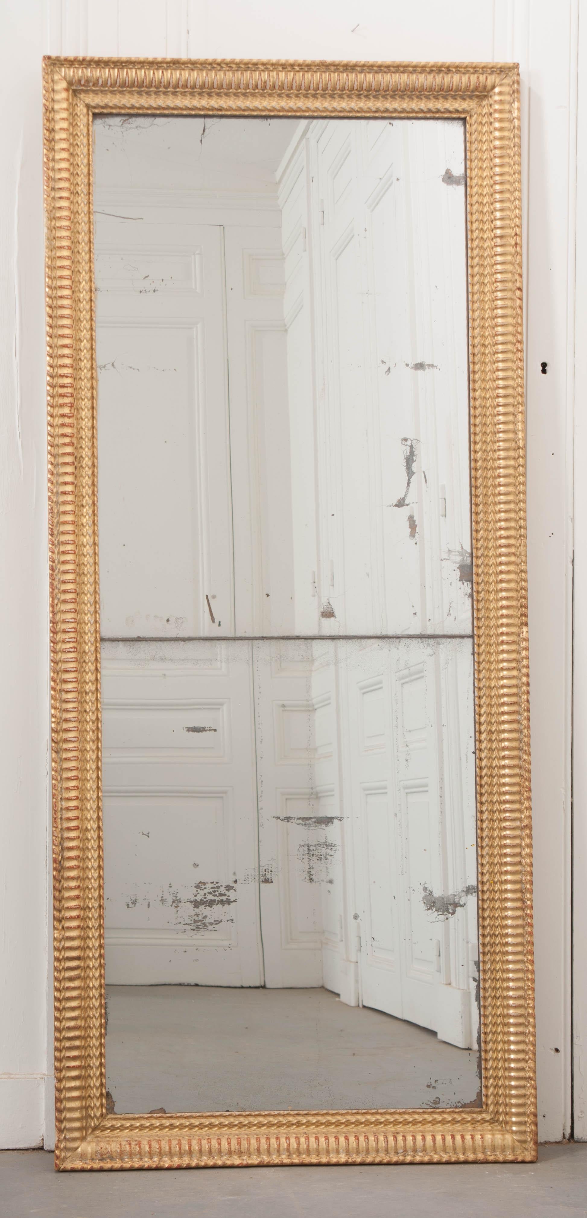 A tall and narrow gold gilt mirror from 19th century France. The mirror’s rectangular frame has a special design, with ribbed and running-chevron motifs finished in brilliant gold gilt. The frame has some French red that is visible from behind the