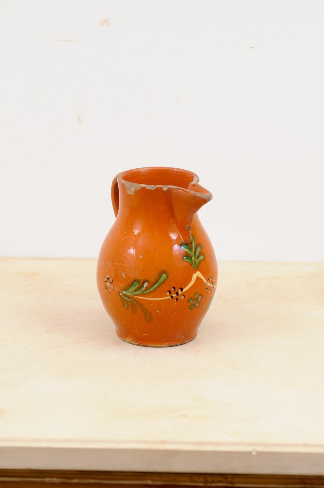 A French redware pitcher from the 19th century, with orange, cream and green glaze and floral motifs. Created in France during the 19th century, this redware pitcher charms us with its vivid colors and stylized floral motifs. Presenting a handle in
