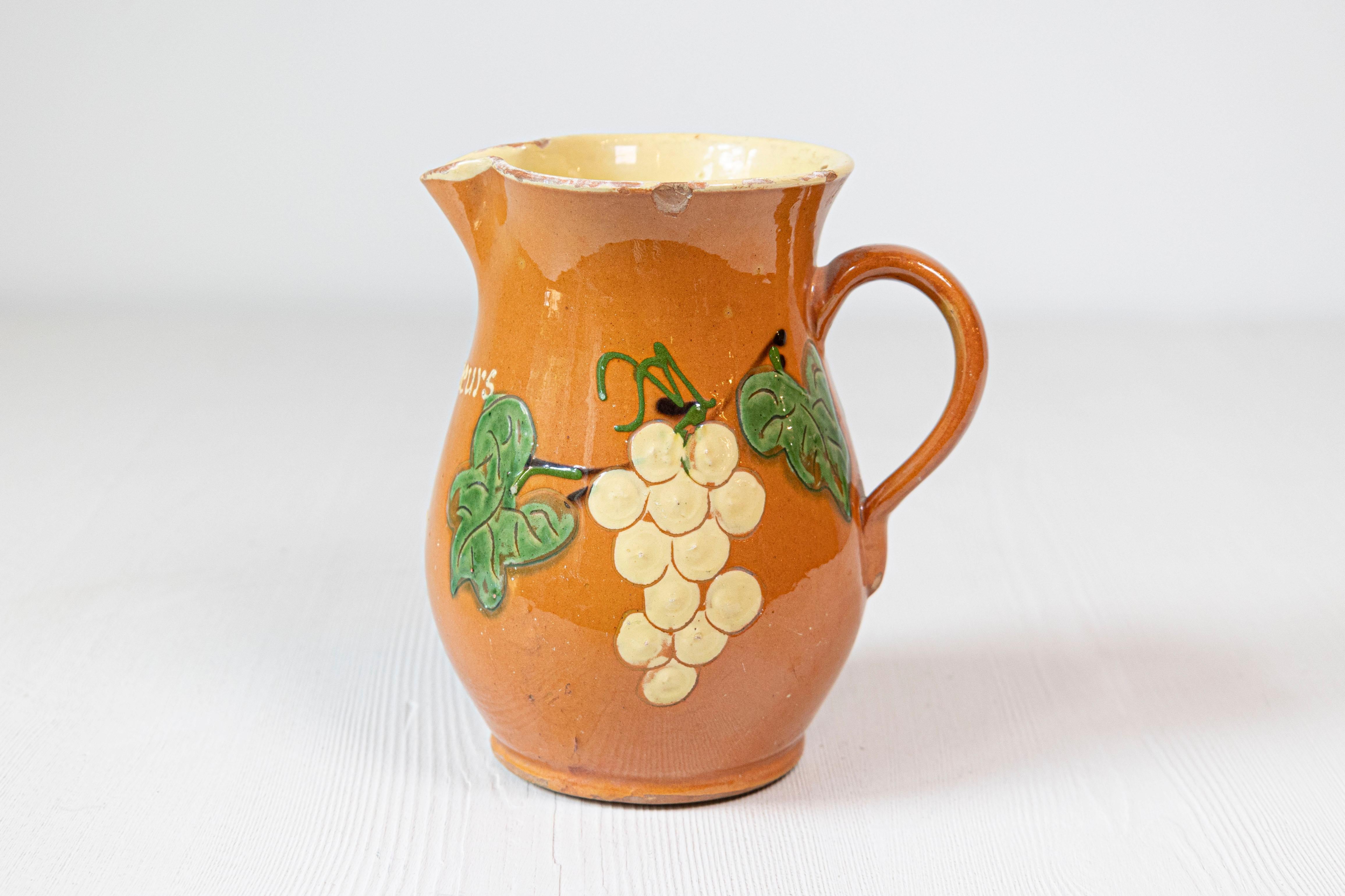 Una jarra de vino de loza roja francesa del siglo XIX, con vidriado naranja, crema y verde y motivos de uvas. Creadas en Francia durante el siglo XIX, cada una de esta pareja de jarras de loza roja nos encanta por sus vivos colores perfectamente
