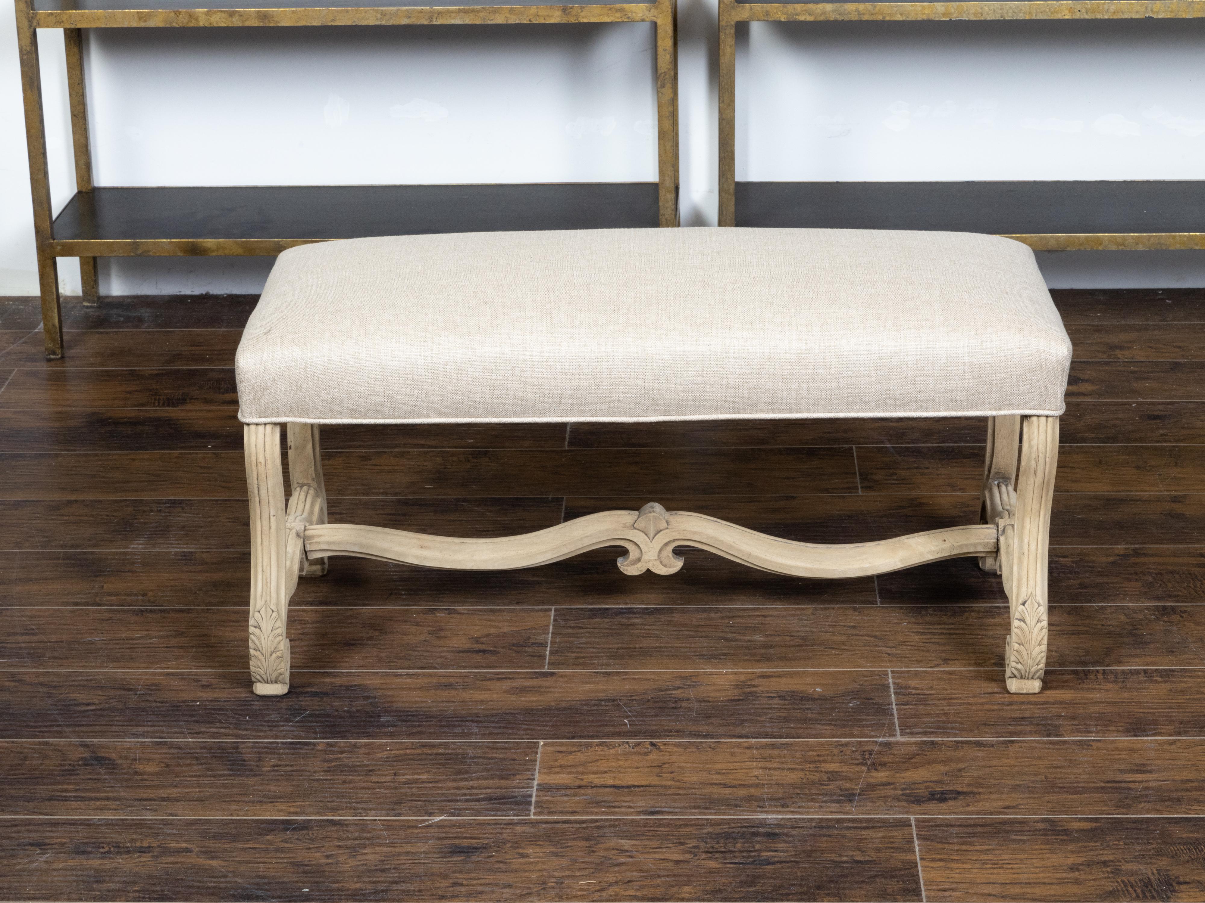 A French Rococo style bleached wood bench from the 19th century, with new custom linen upholstery, curving legs, carved acanthus leaves and H-form cross stretcher. Created in France during the 19th century, this bench features a Rococo style