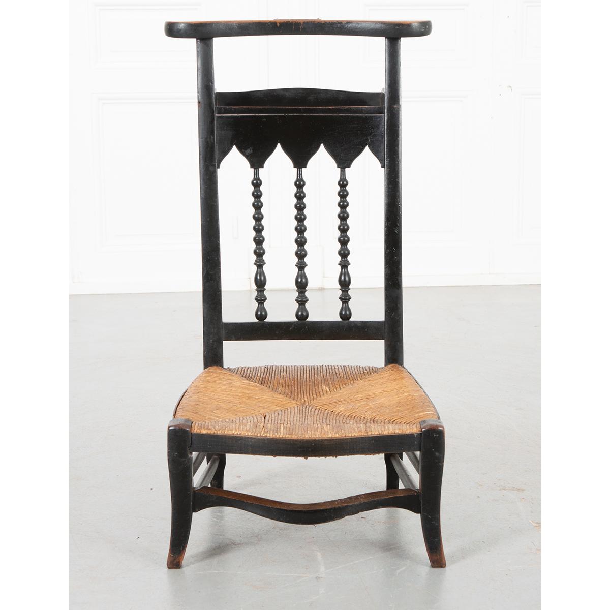 This is a French 19th century rush seat prie dieu. It has a painted wood finish with turned wood spindles on the back and also a shelf. It has a shaped wood top where your arms would have rested for praying. A brass plaque rests on top with the name