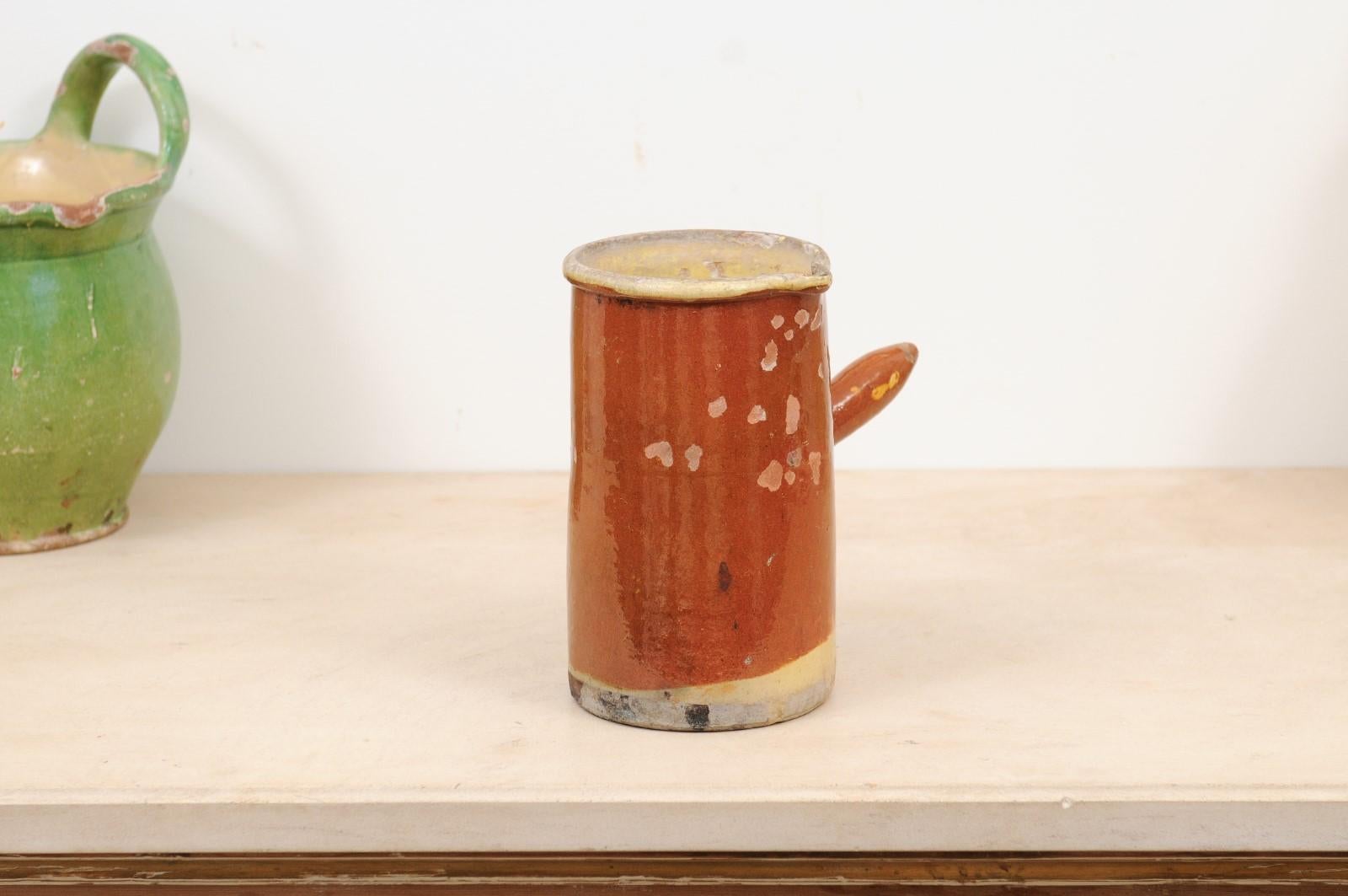 A French rustic pitcher from the 19th century, with rust colored glaze and straight handle. Created in France during the 19th century, this pitcher features a cylindrical body adorned with a rust ground showing a nicely weathered appearance.