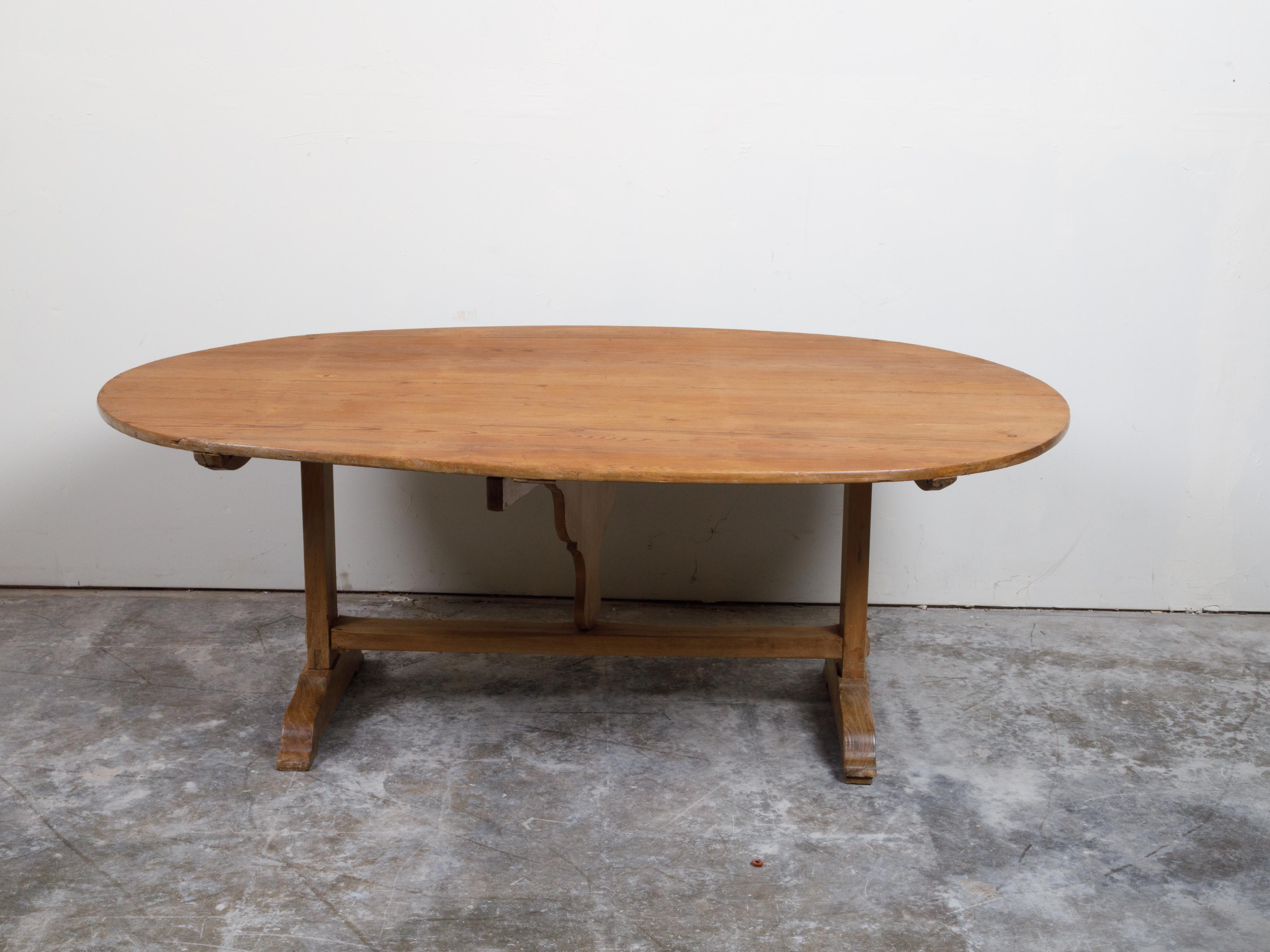 A French large walnut tilt-top table from the 19th century, with oval top and trestle base. Created in France during the 19th century, this large walnut farm table features an oval tilt top resting above a rustic trestle base with simple wedge. With