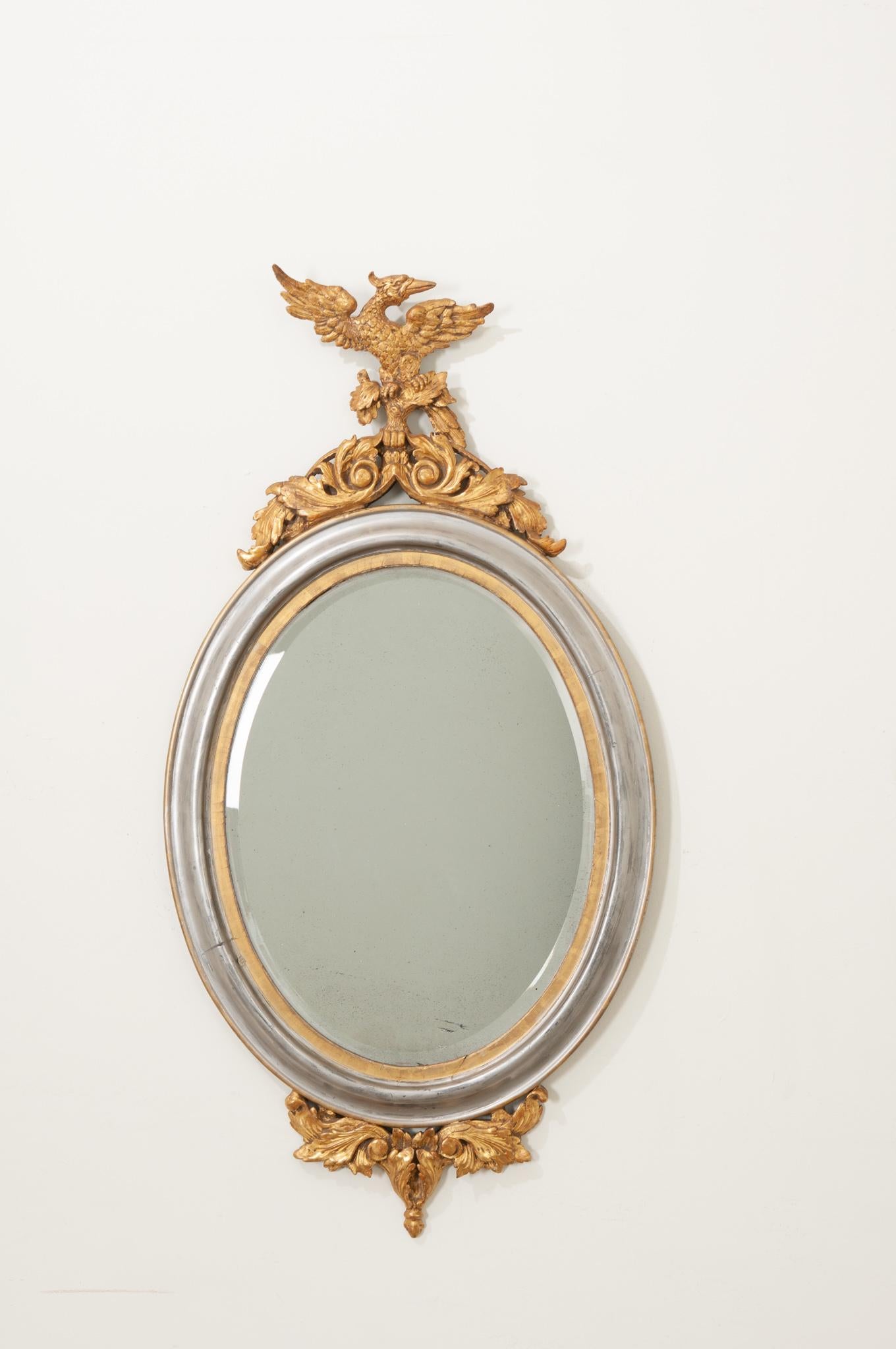 A regal gold and silver gilt mirror with intricate, carved details at the top and bottom. A splendid set of Ho ho birds top scrolling acanthus leaves on the crown of an elegant oval frame that terminates in a snake emerging from acanthus supports.