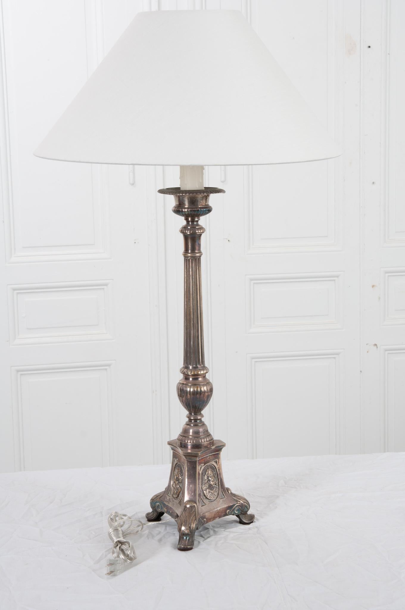 The base of this lamp is a silver plated 19th century candlestick from France. Most likely used on an altar in a Christan church, evident from the images of the Holy Trinity on the heavy tripod base. Beautiful details throughout the candlestick