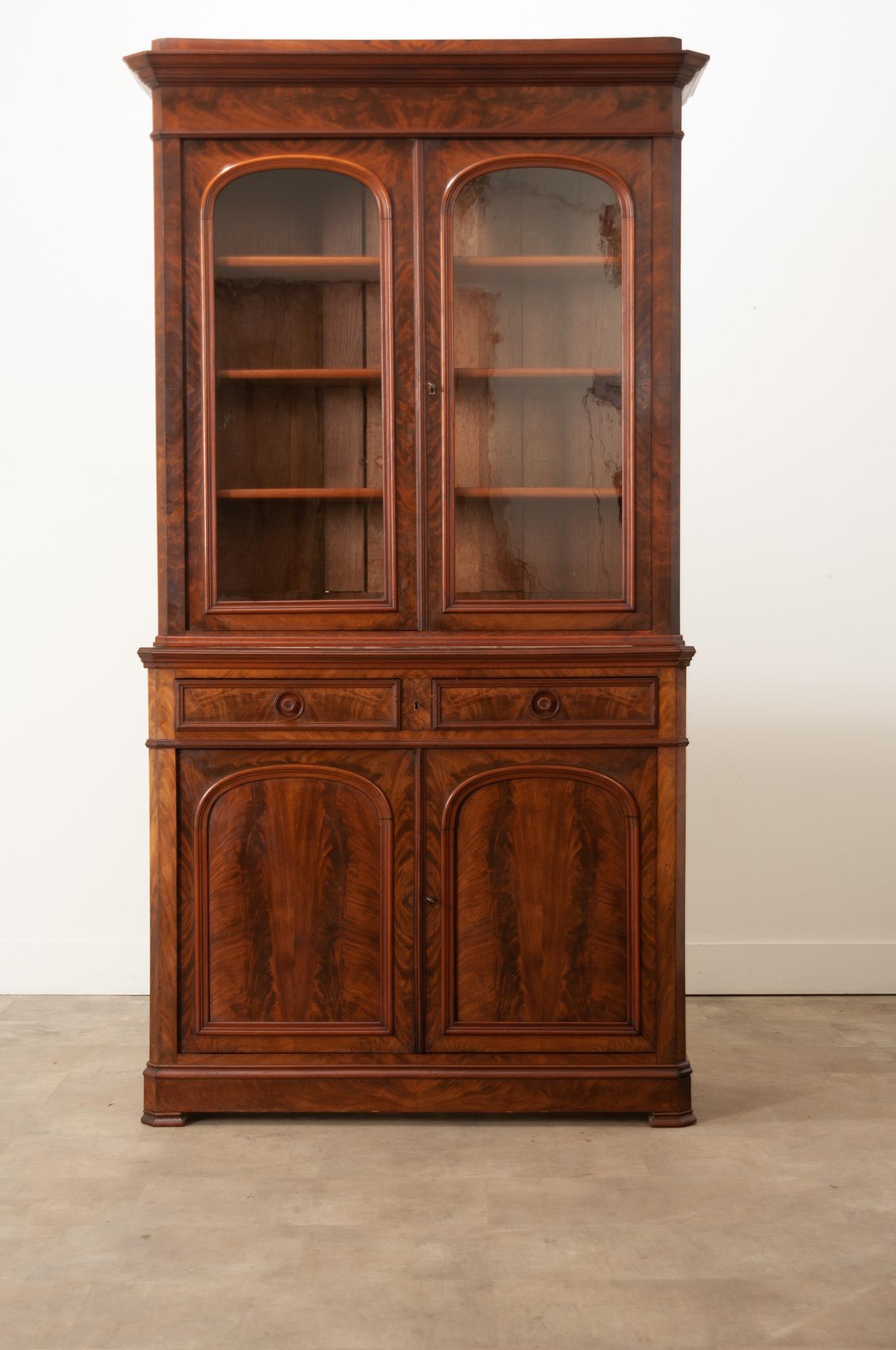 This handsome bibliotheque was crafted in the style of Louis Philippe in 19th century France. Gorgeous flame patterned mahogany is well paired with deep red toned trim to create a knockout case piece. Original wavy glass in the top cabinet doors