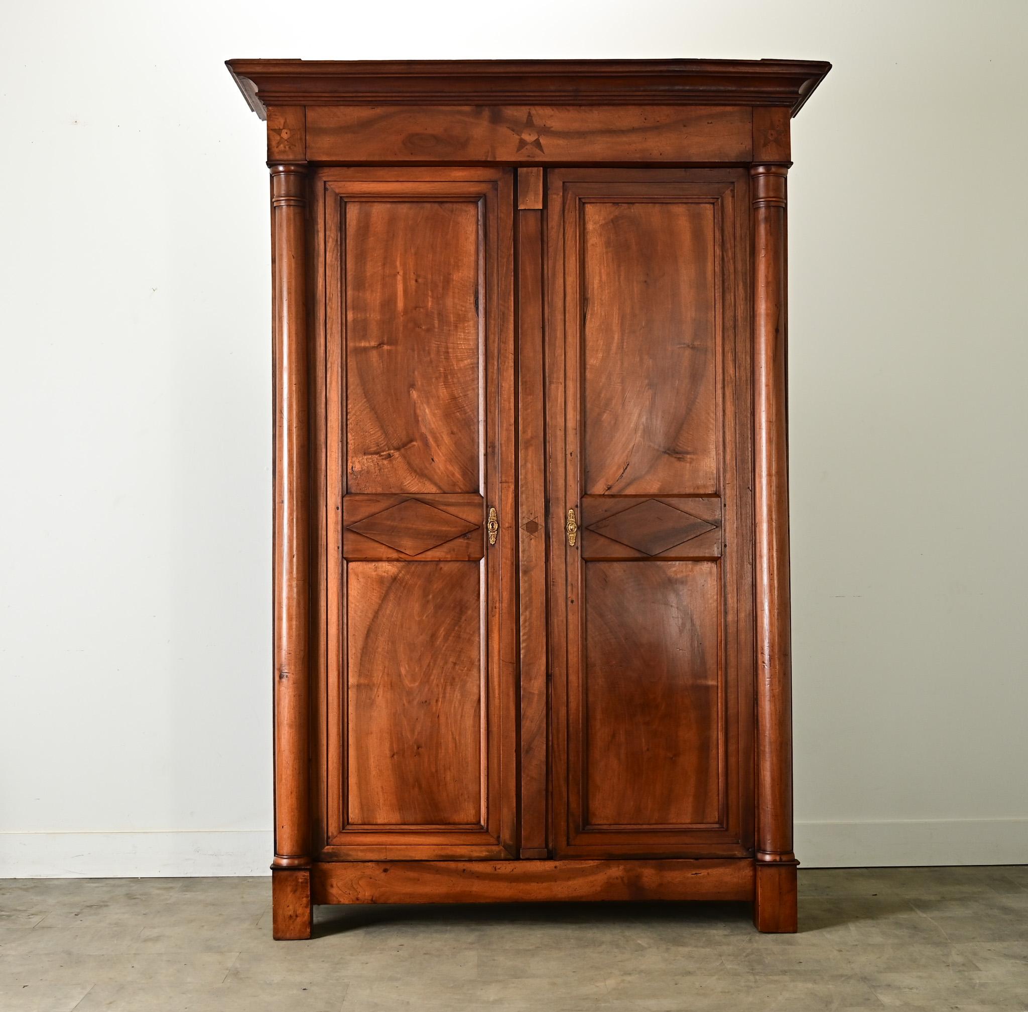A towering French Empire armoire from the late 1800s, made of beautifully figured walnut. The pronounced crown hangs over a frieze decorated with a marquetry star above each ½ round pilaster on the sides and one star at the center. Each paneled and