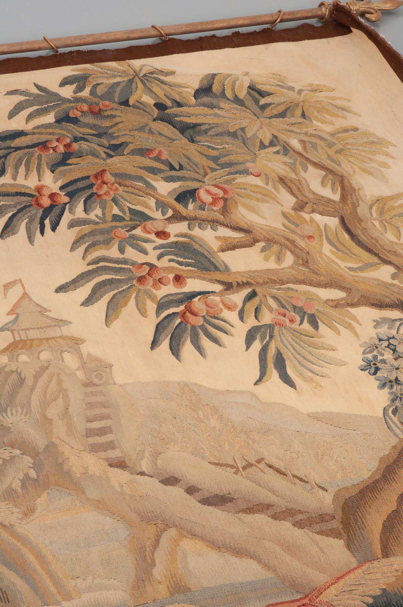 This 19th century French Tapisserie de Verdure, circa 1850s, is exquisite. It is wool and cotton, and hangs on a metal tapestry rod with rings. The scene is framed by beautiful trees and depicts most prominently a pagoda and exotic birds in the