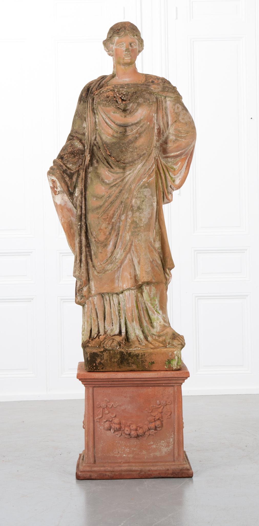 This patinated terracotta statue rests atop a newer pedestal that would work beautifully indoors or outdoors. It depicts a robed female with one hand tucked behind her back and the other hand is missing. The dimensions of the statue alone are: 61” H