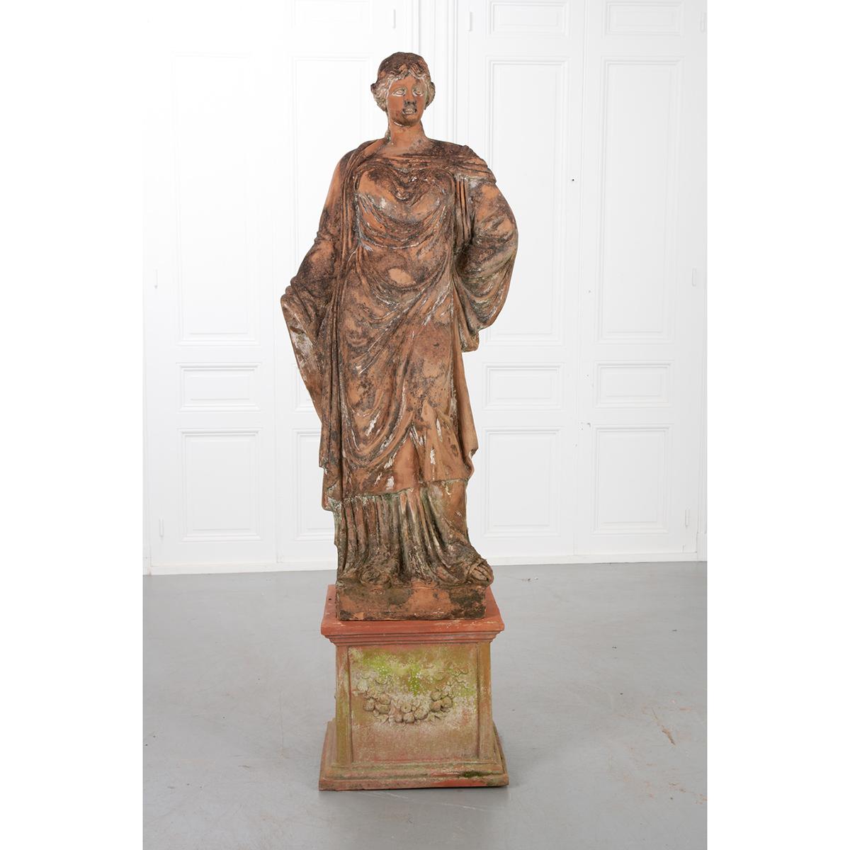 This patinated terracotta statue rests atop a newer pedestal that would work beautifully indoors or outdoors. It depicts a robed female with one hand tucked behind her back and the other hand is missing. The dimensions of the statue alone are: