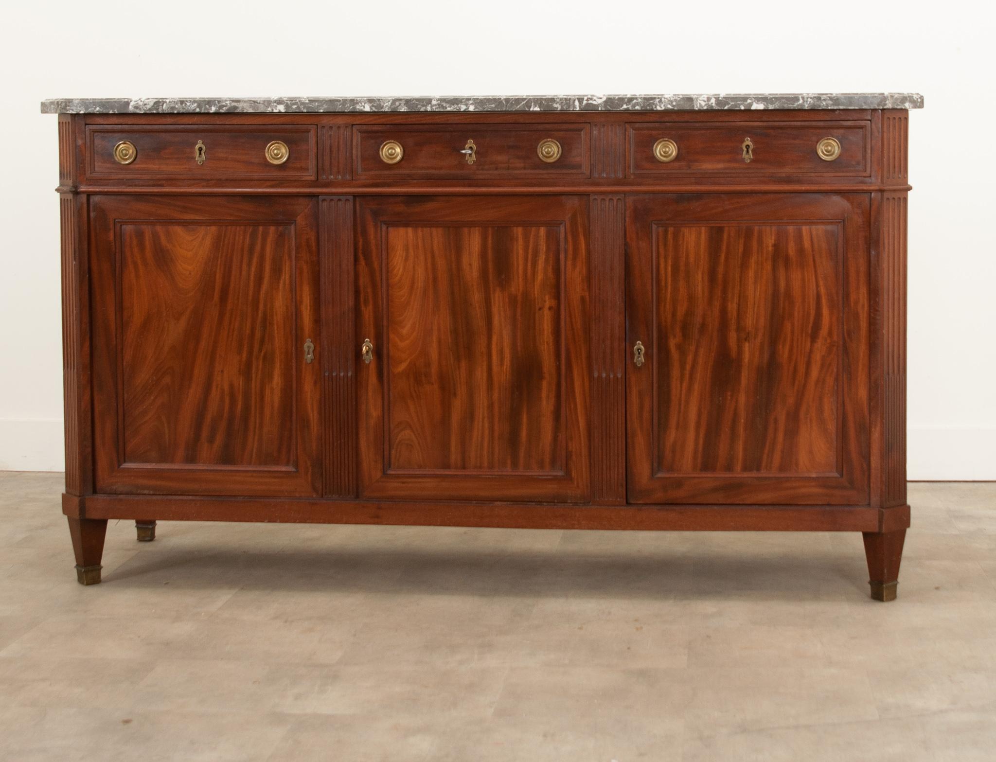 Fabulous fluted detailing and flame mahogany make this 19th century transitional French mahogany enfilade truly stunning. This solid mahogany case piece incorporates elements of both Louis XVI and Directoire styles. A shaped charcoal and gray marble