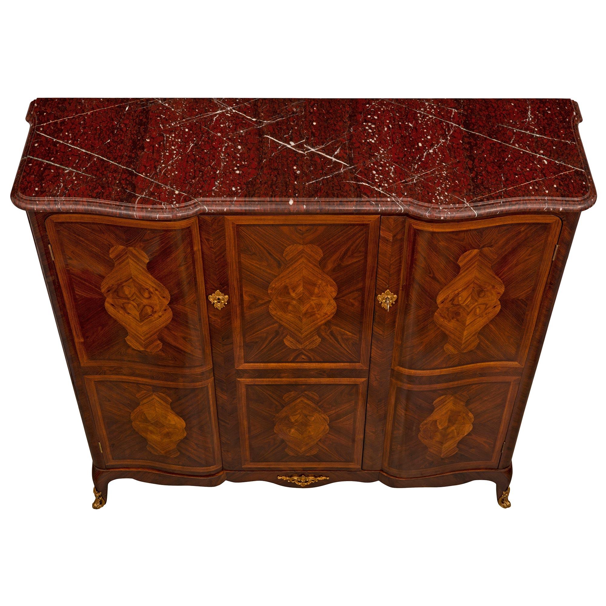 An elegant French 19th century Louis XV st. Kingwood, Tulipwood, ormolu, and Rouge Griotte marble cabinet. The three door cabinet à hauteur d'appui is raised by tapered lightly curved legs with fine pierced foliate ormolu sabots below the scallop
