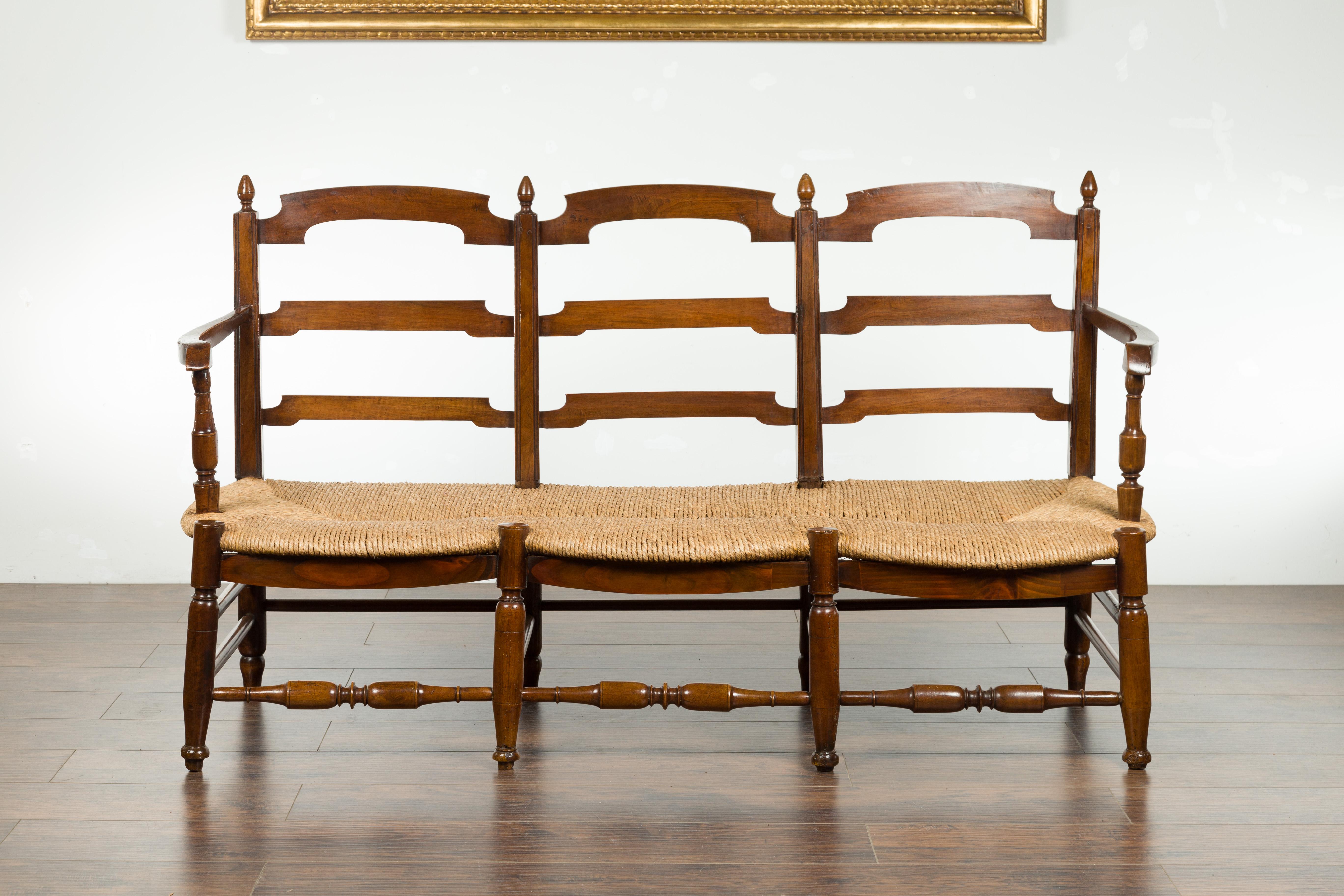 A French walnut bench from the 19th century, with open back and rush seat. Created in France during the 19th century, this walnut bench features an open ladder style back, connected to two arms with turned supports, resting on a long rectangular