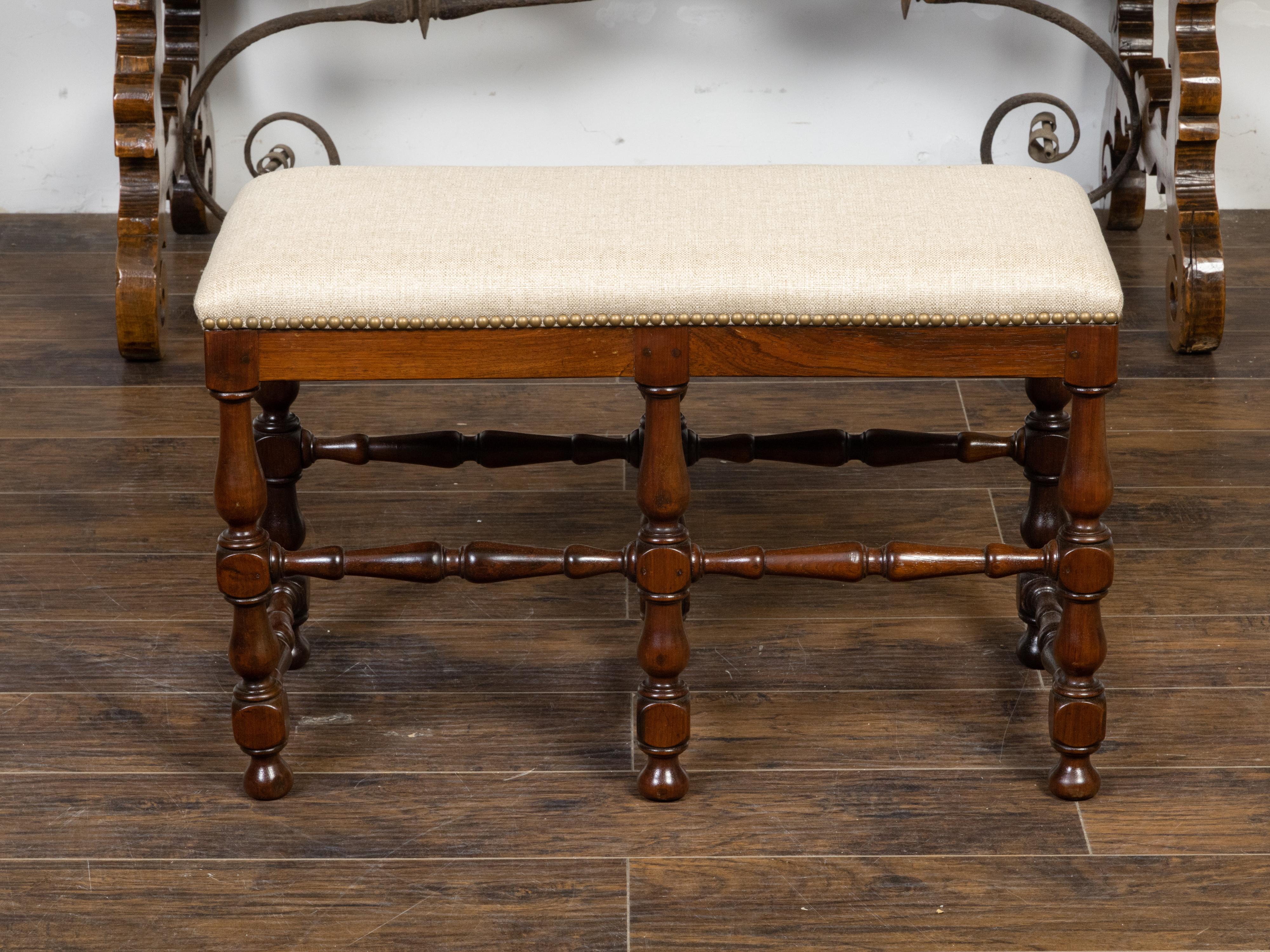 A French walnut bench from the 19th century with new linen upholstery, brass nailhead trim, turned baluster legs and cross stretchers. Created in France during the 19th century, this walnut bench features a rectangular seat newly recovered with a