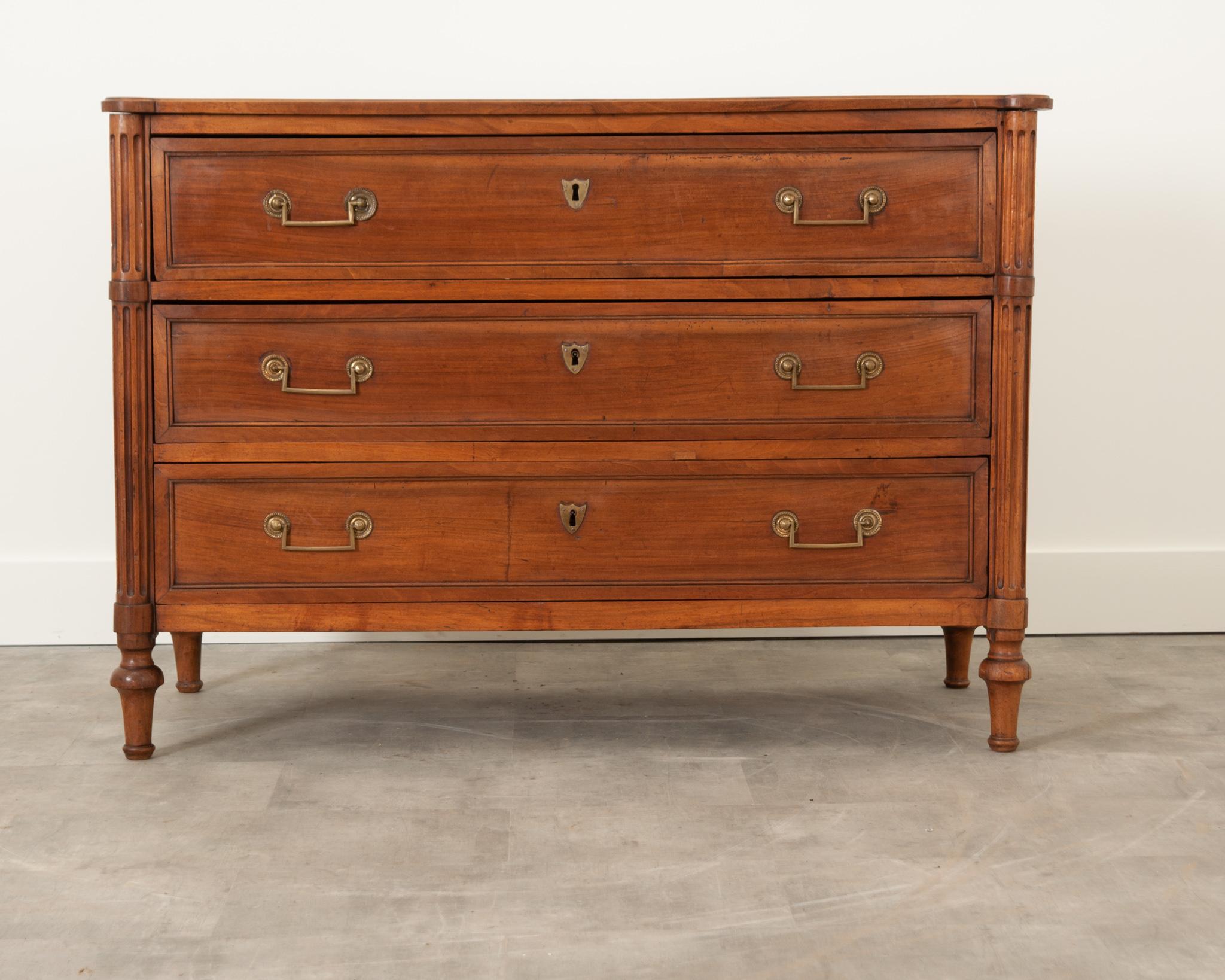 A handsome solid walnut commode from 19th century France, crafted in the style of Louis XVI. The three sizable paneled drawers are flanked by tureted, fluted corners. Each drawer is fixed with a pair of cast brass bail pulls and a shield shaped