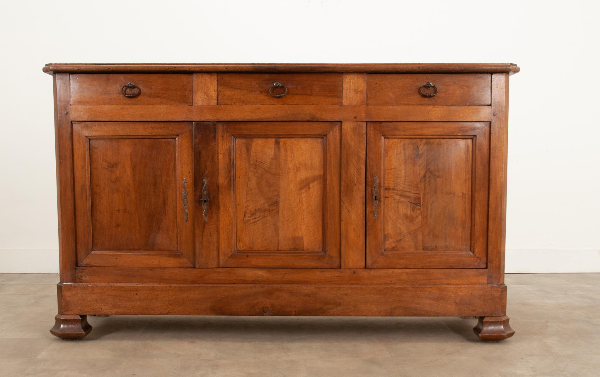 This attractive Louis Philippe style solid walnut enfilade with canted corners, c. 1870, features a gorgeous patinated finish highlighting the rustic charm of this piece. The wood top sits above an apron with three drawers, each with an oval brass