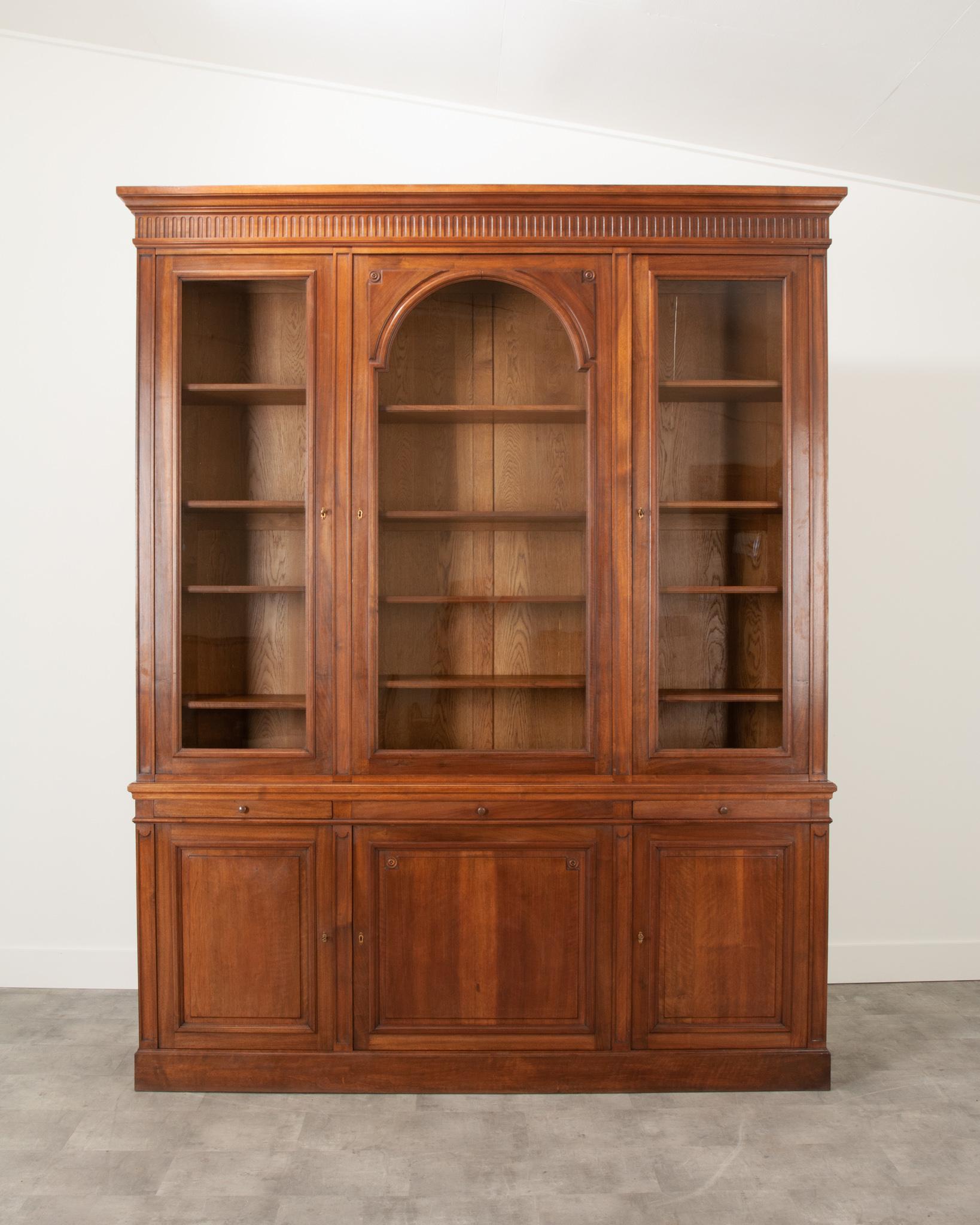 A massive French walnut Bibliotheque or bookcase from the 19th Century. The straight lines and vertical motif across the crown are classic decorations of the Louis XVI style. All three of the glass door fronts contain the original wavy glass, the