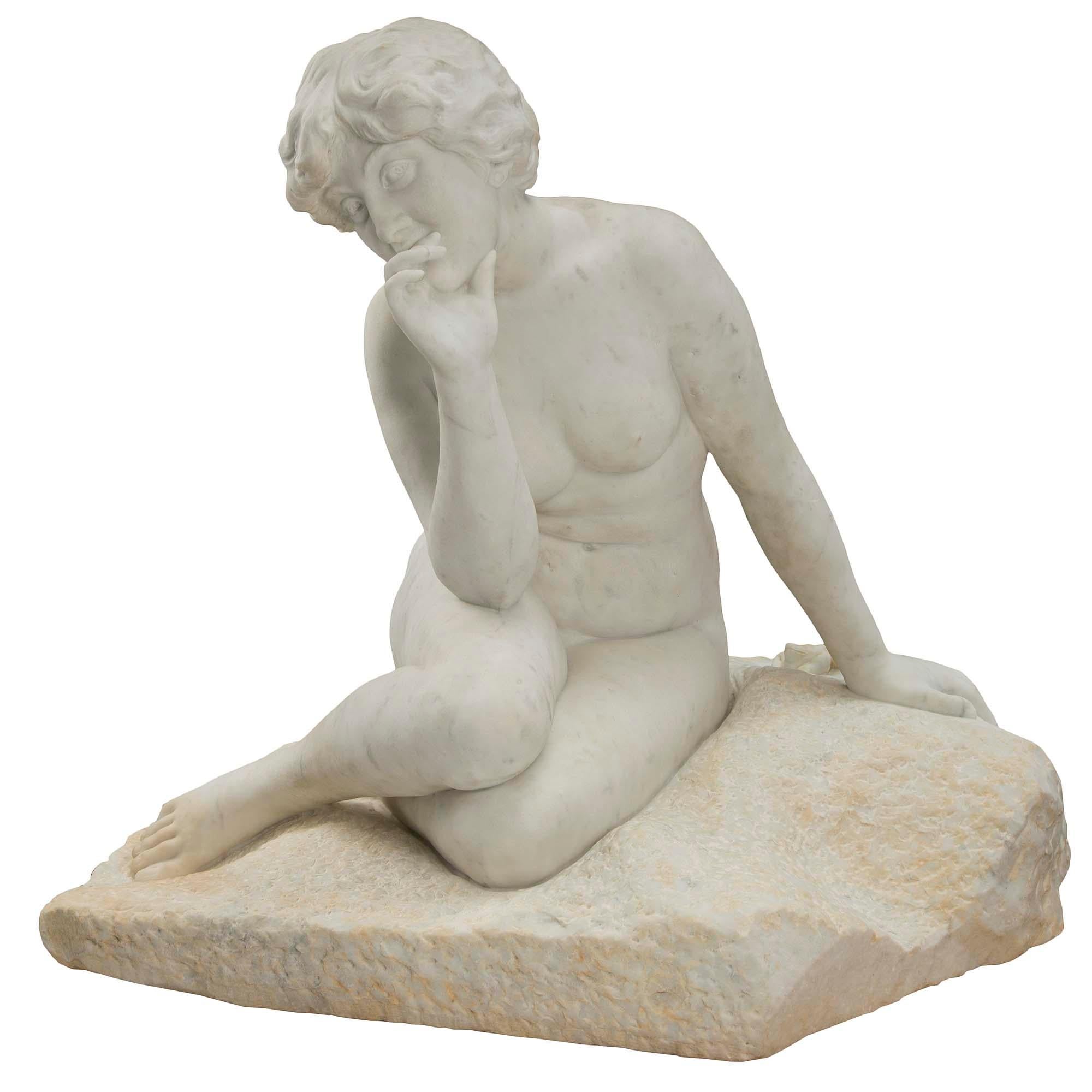 A wonderful French 19th century white Carrara marble statue of a seated young lady. The charming statue is raised by a richly sculpted rocky base where the beautiful nude woman sits elegantly with her hand at her face with a pensive air. One would