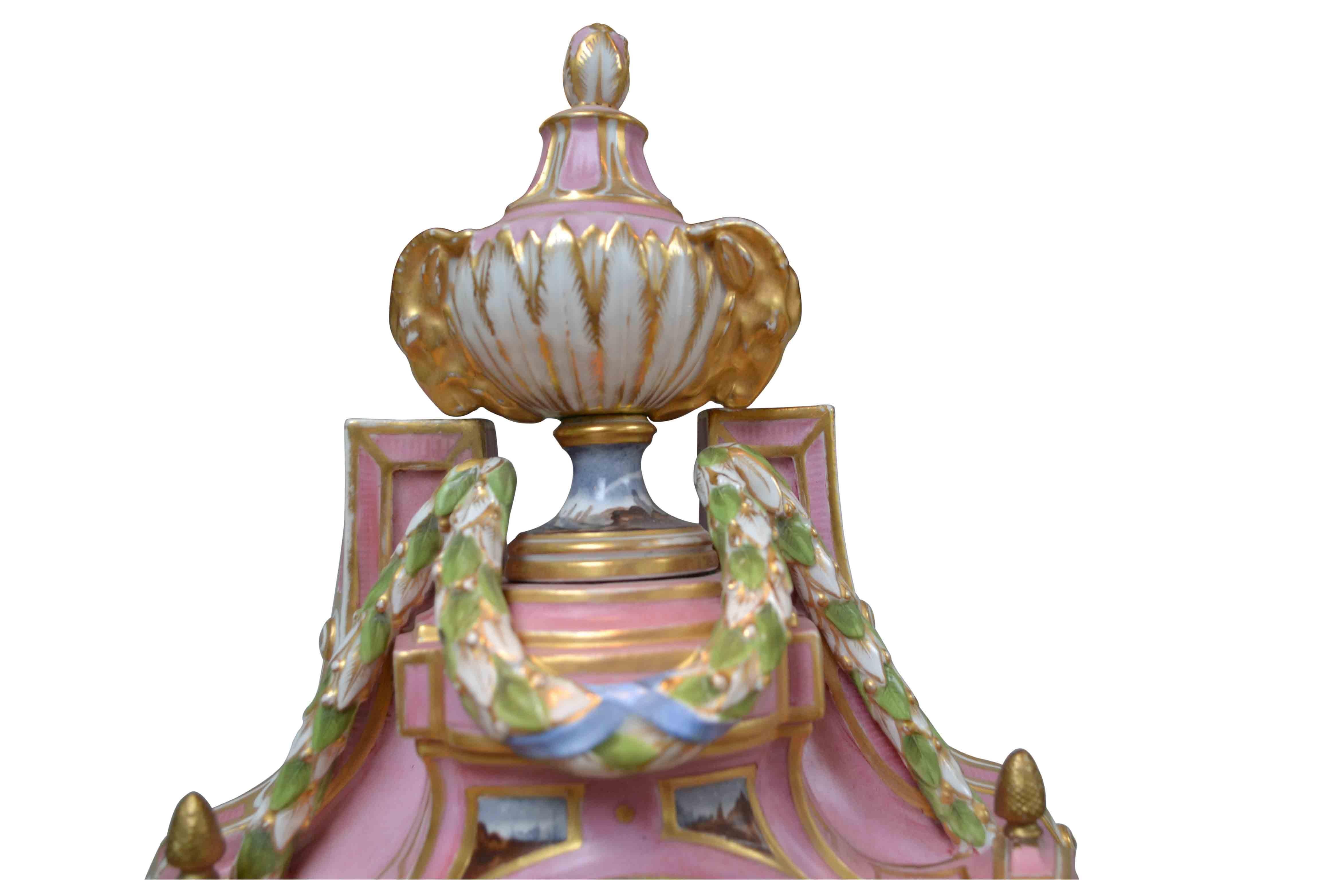 A finely decorated pink porcelain Cartel clock in the Louis XVI style; the predominantly pink case surmounted by a classical urn with goats heads and colored enamel swags. Below the white enamel dial with Roman numerals is a finely painted village