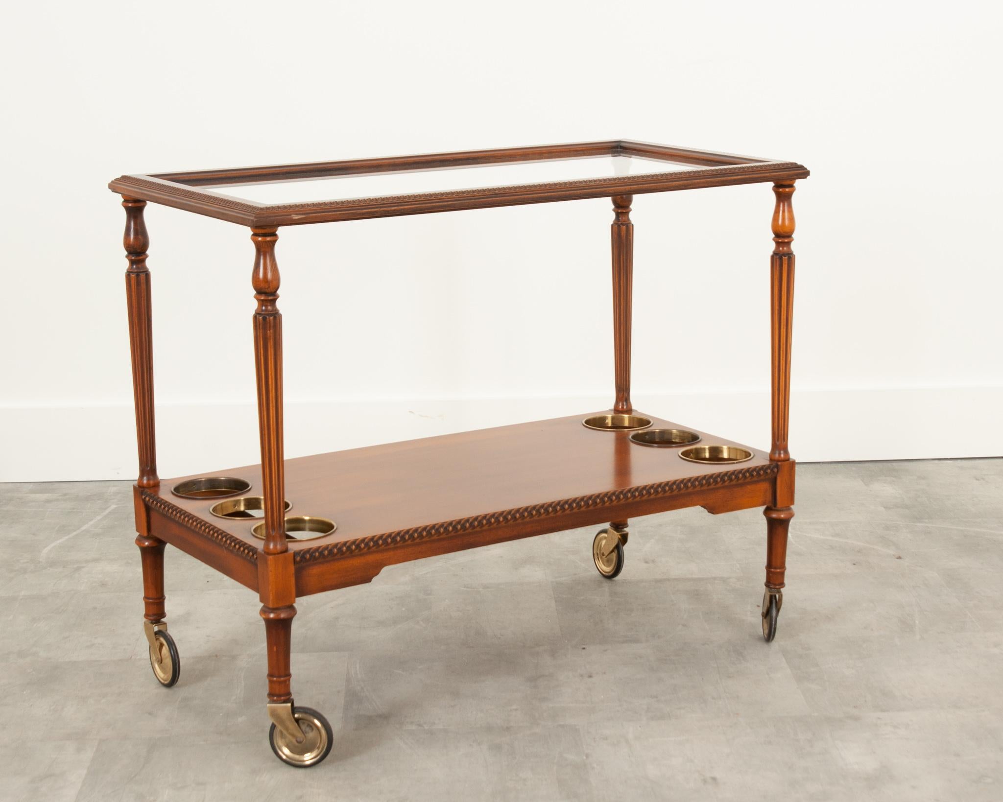 This vintage French bar cart is the perfect antique element to elevate any bar room or living space. The warmly toned mahogany frame suspends the glass top and showcases decorative carving around the perimeter. Turned, fluted legs connect to a lower