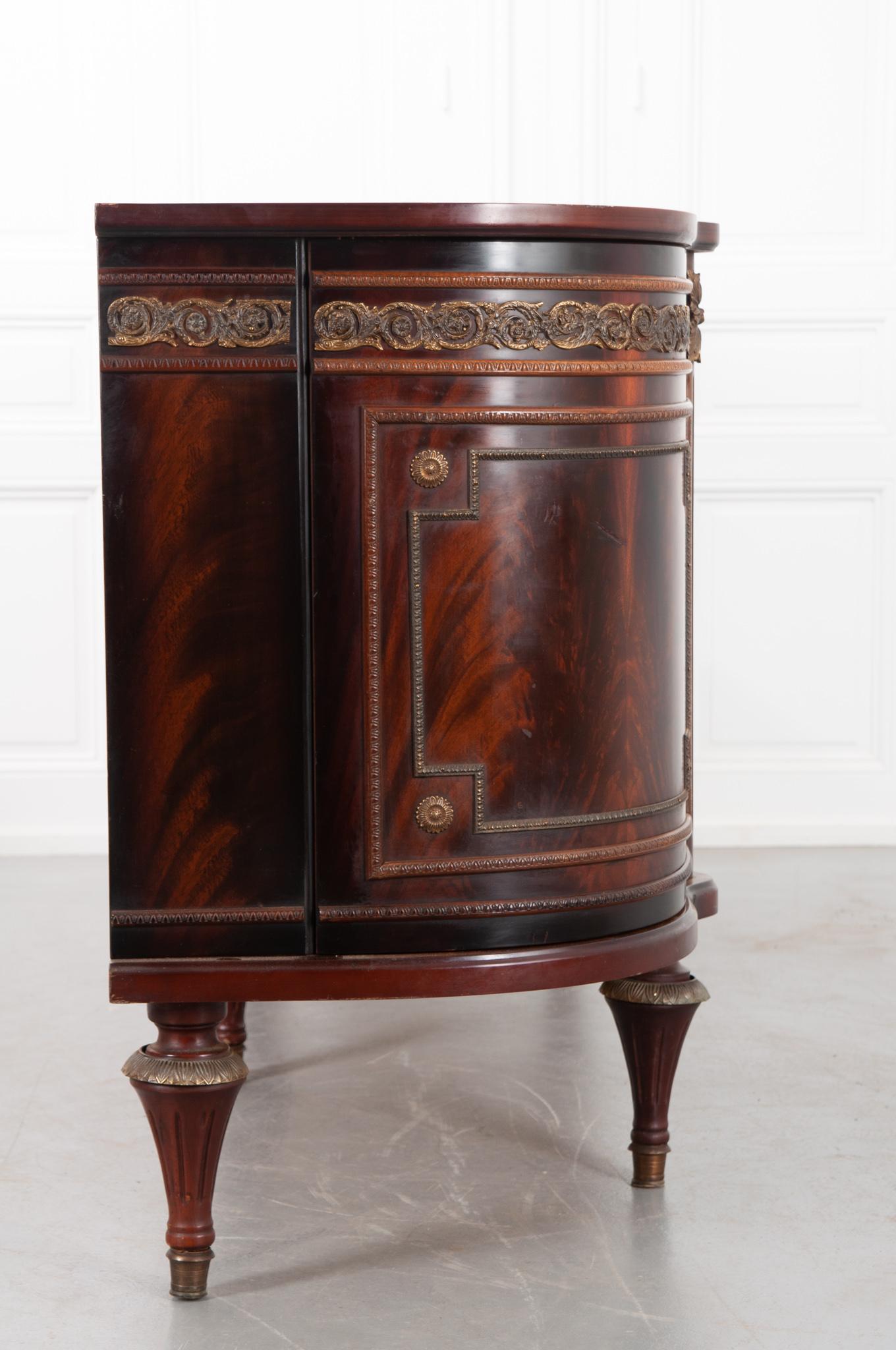 This is a French 20th century five door demilune enfilade, made of mahogany veneer with impressive ormolu accents. The curved end doors conceal two fixed glass shelves each. The center doors allow for more storage space with the far left door and