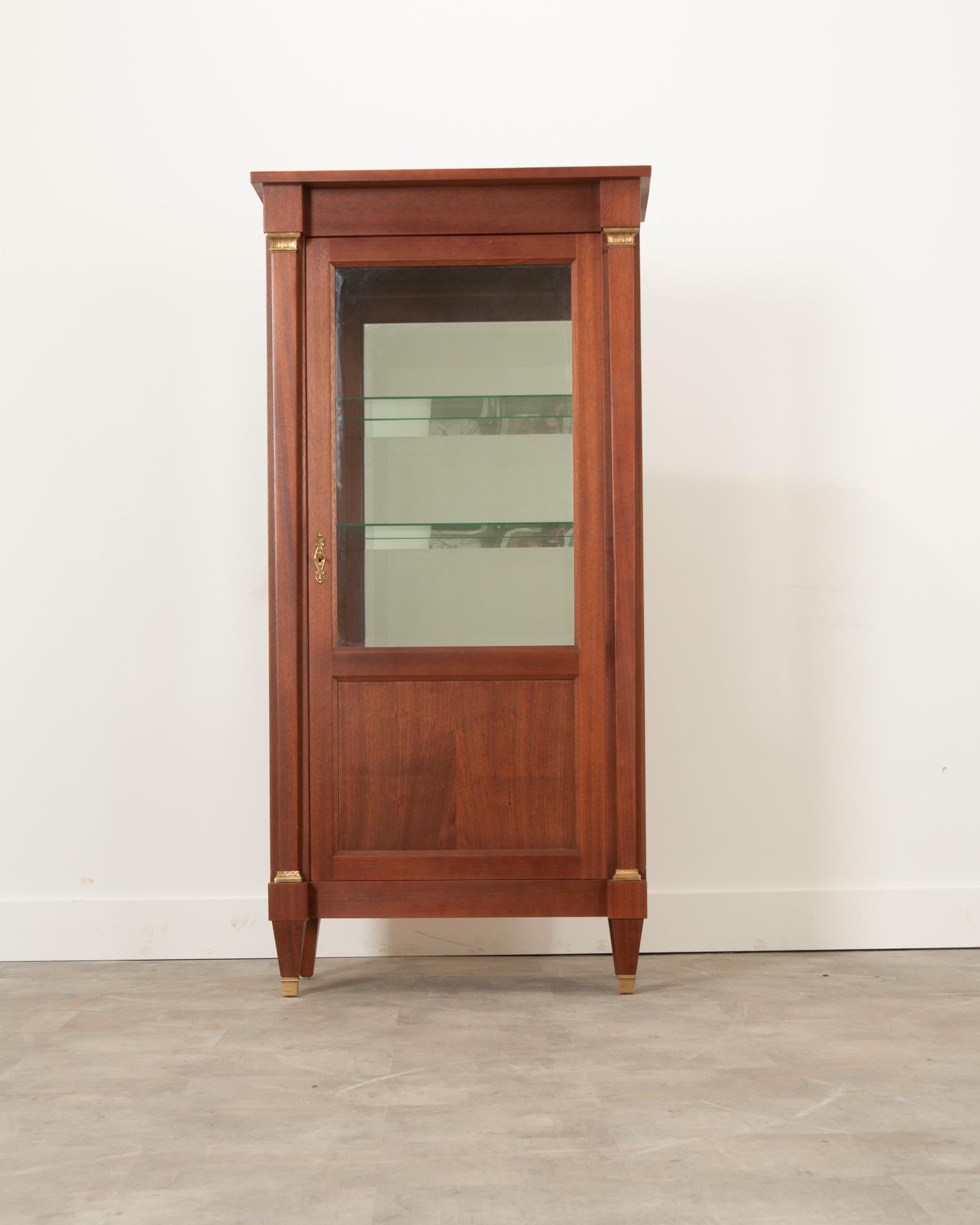 Display your treasured objects in this beautiful early 20th century French Empire style mahogany vitrine. Beveled glass panels can be found in the case antique’s door and sides, allowing one to view the contents placed inside on its two glass