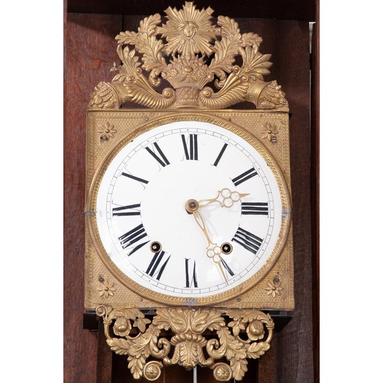 A carved crest of a basket with flowers crowns this heavily carved oak case clock, c. mid 1800’s.  A glazed-panel door just below the crest is flanked by sides with pierced half-moon carvings and houses the painted enamel clock face with Roman