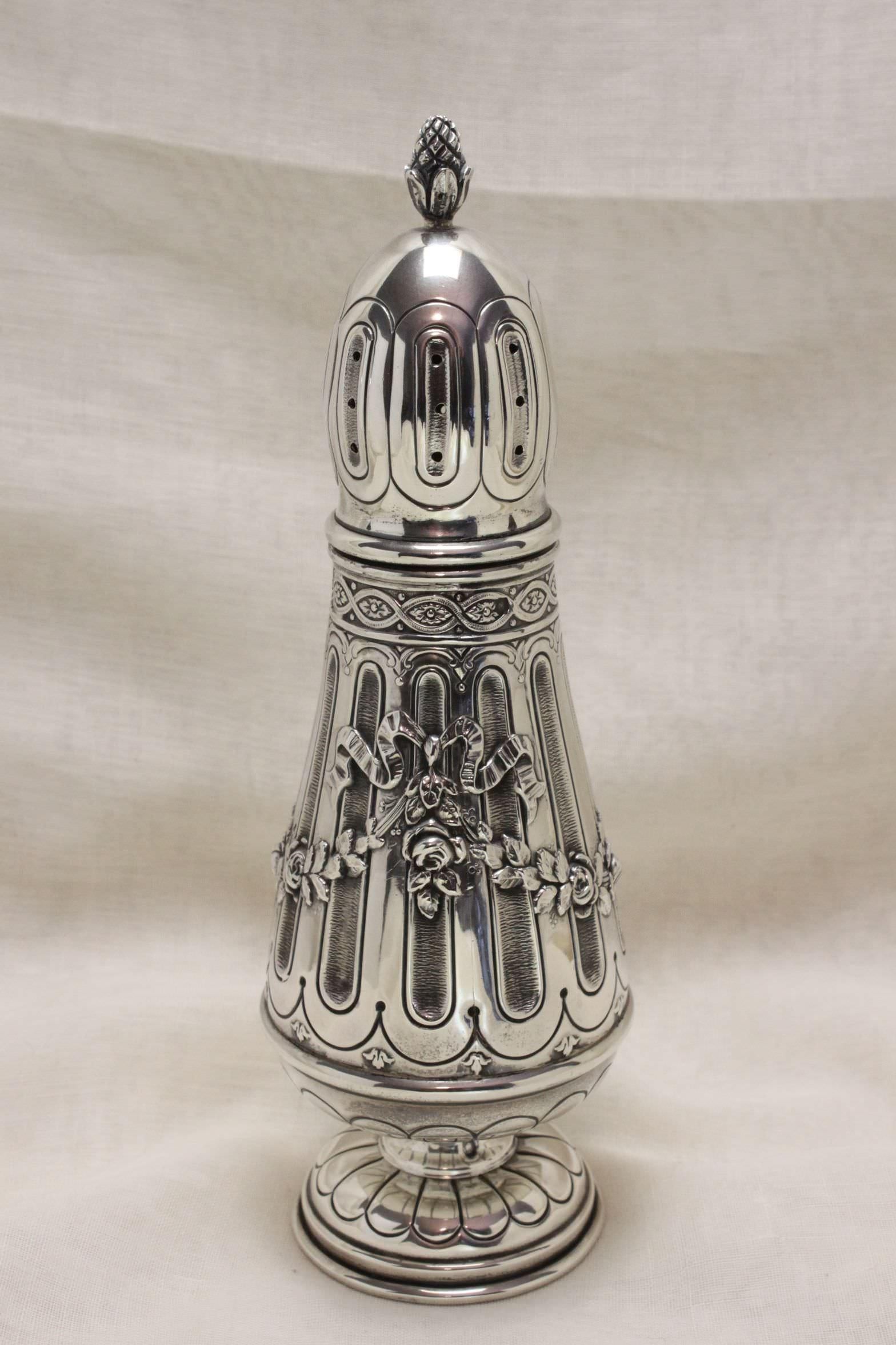 The fluted body of this very pretty sugar caster is decorated with embossed ribbons and swags of roses and stands on an embossed circular foot. The domed lid also features fluting and is topped with a pineapple. The French stamped mark of Minerva's