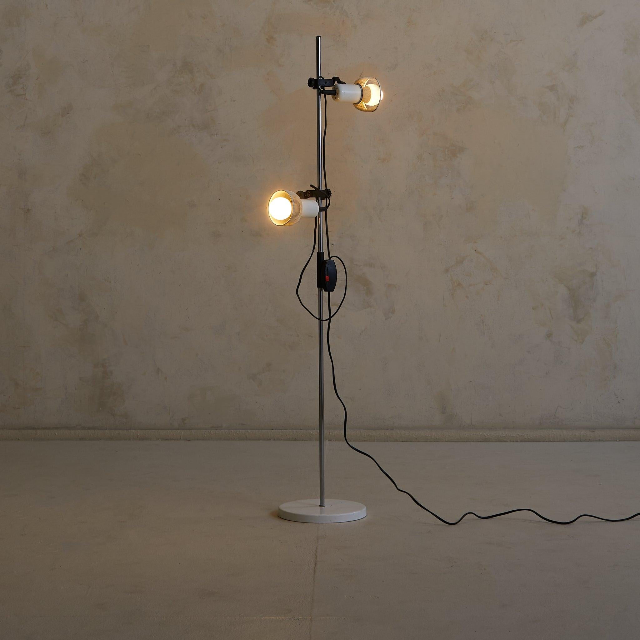 Questa lampada da terra francese di metà secolo incarna l'abbraccio dell'epoca al minimalismo industriale. Due paralumi in metallo traforato, rifiniti in una morbida tonalità avorio, sono sospesi da un sottile stelo cromato in cima a una base