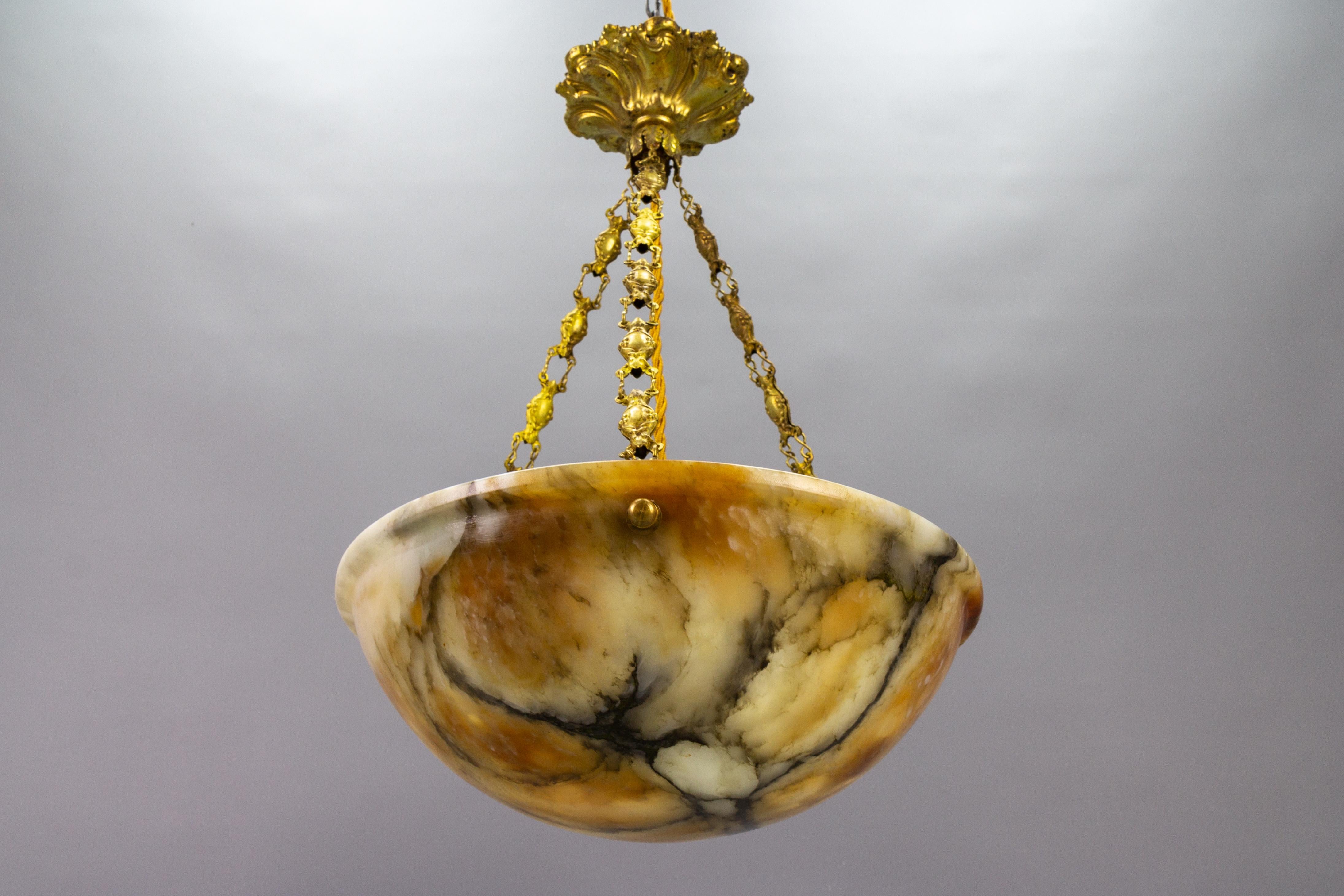 An adorable French pendant light featuring an alabaster shade in a lovely, light and a dark, orange, and cream hue with black veining. The lampshade - bowl is suspended from three ornate brass chains. When lit, alabaster emanates a warm, sun-like