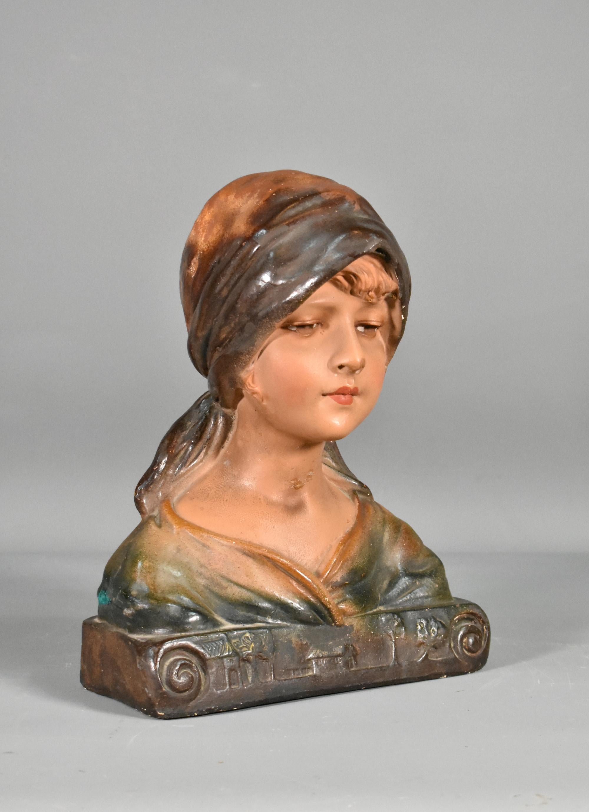 French Antique Bust of a Young Girl in Plaster 

A pretty polychrome plaster bust of a young girl wearing a headscarf with her eyes cast downwards. 

The piece features carvings of buildings and trees across the front base in the Art Nouveau style.