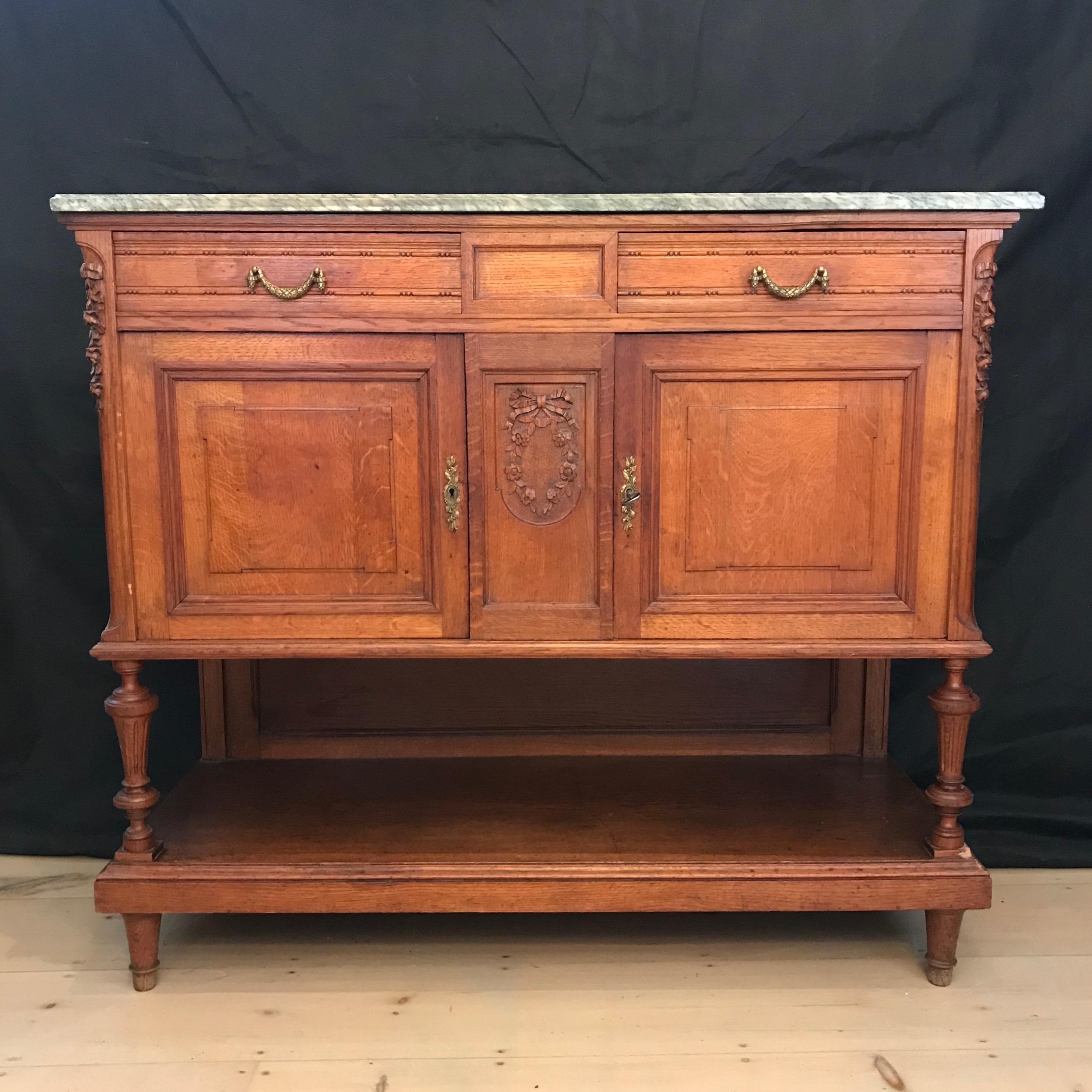This French mid-19th century oak buffet features a grey and white veined marble top with beveled edge and carved ornate corners in the front, over a beautiful honey colored body with great patina. Two drawers with bronze wreathed design are placed