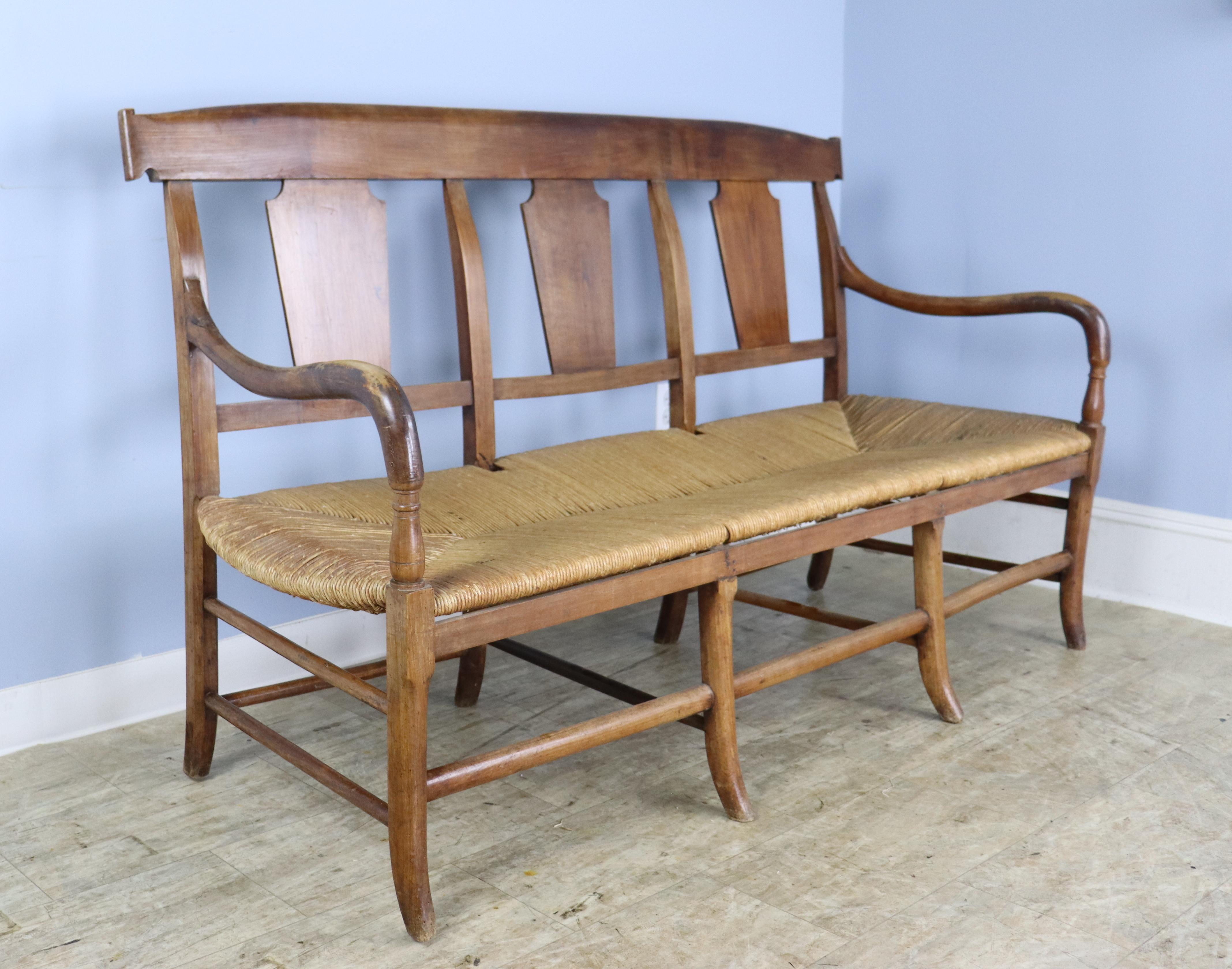 A rush seated French antique bench with Directoire era design elements, specifically the shaped back supports and the carved detial just below the curved arms.  The cherry is a warm brown with good grain and some period appropriate wear, and the