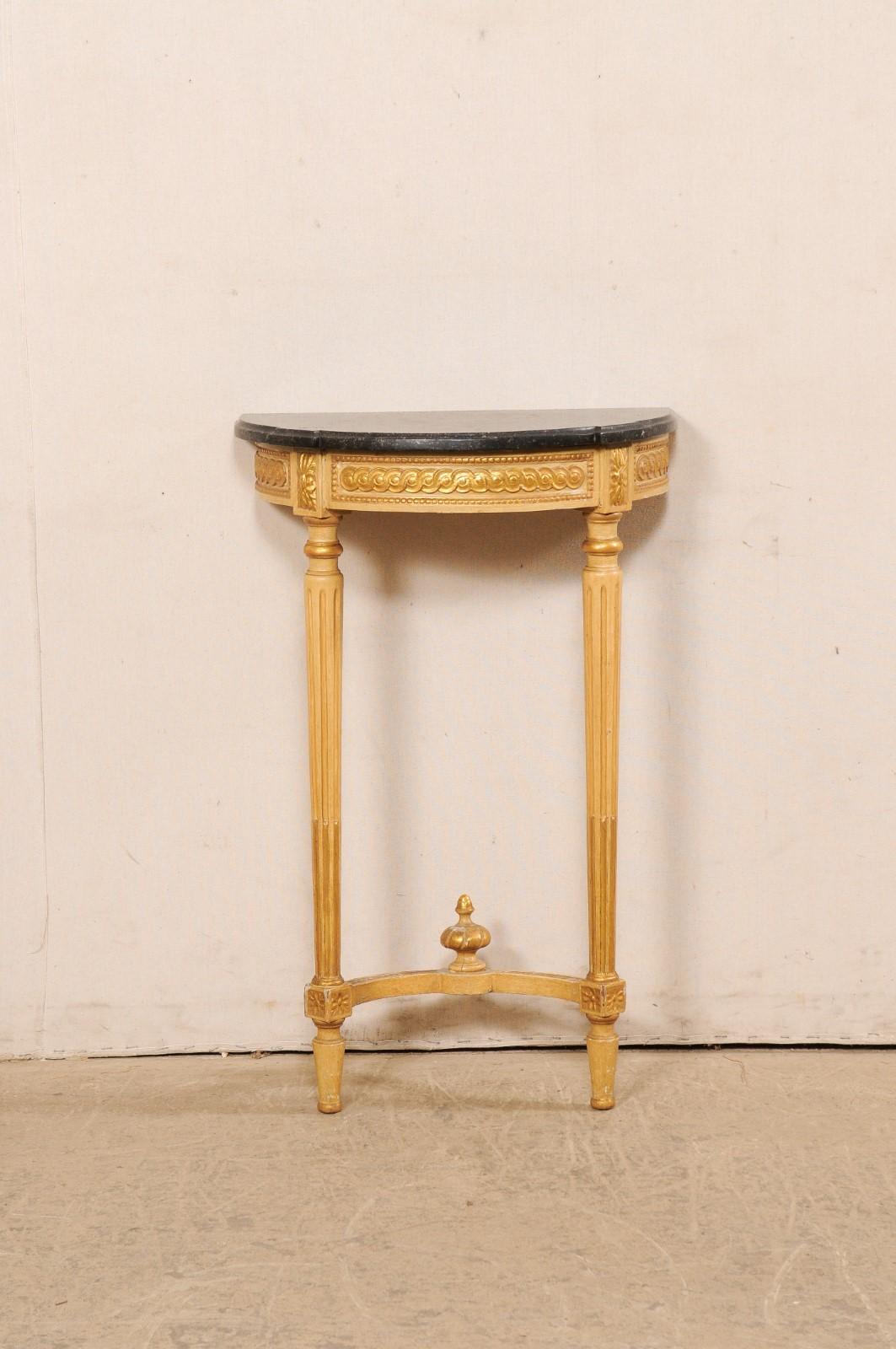 A French small-sized carved and gilt wood wall console, with marble top and finial accent, from the early 20th century. This table from France features a petite demi-shaped (half moon) marble top with squared step-out style corners flanking its