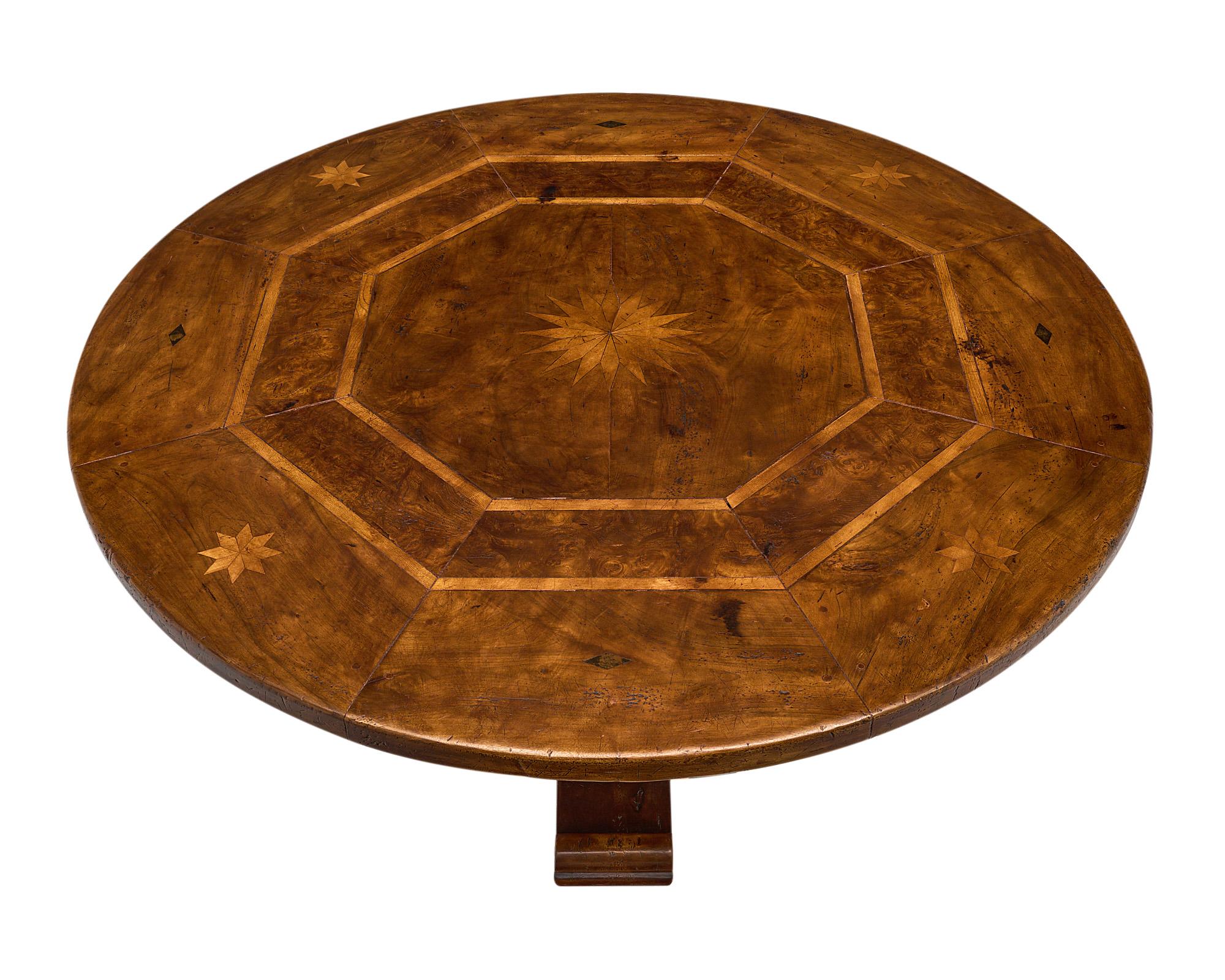 Round dining room table, French, made of solid walnut and burled walnut with inlaid details of a central star. This piece features pegged construction, a hand carved central pedestal, and four scrolled feet. 
 