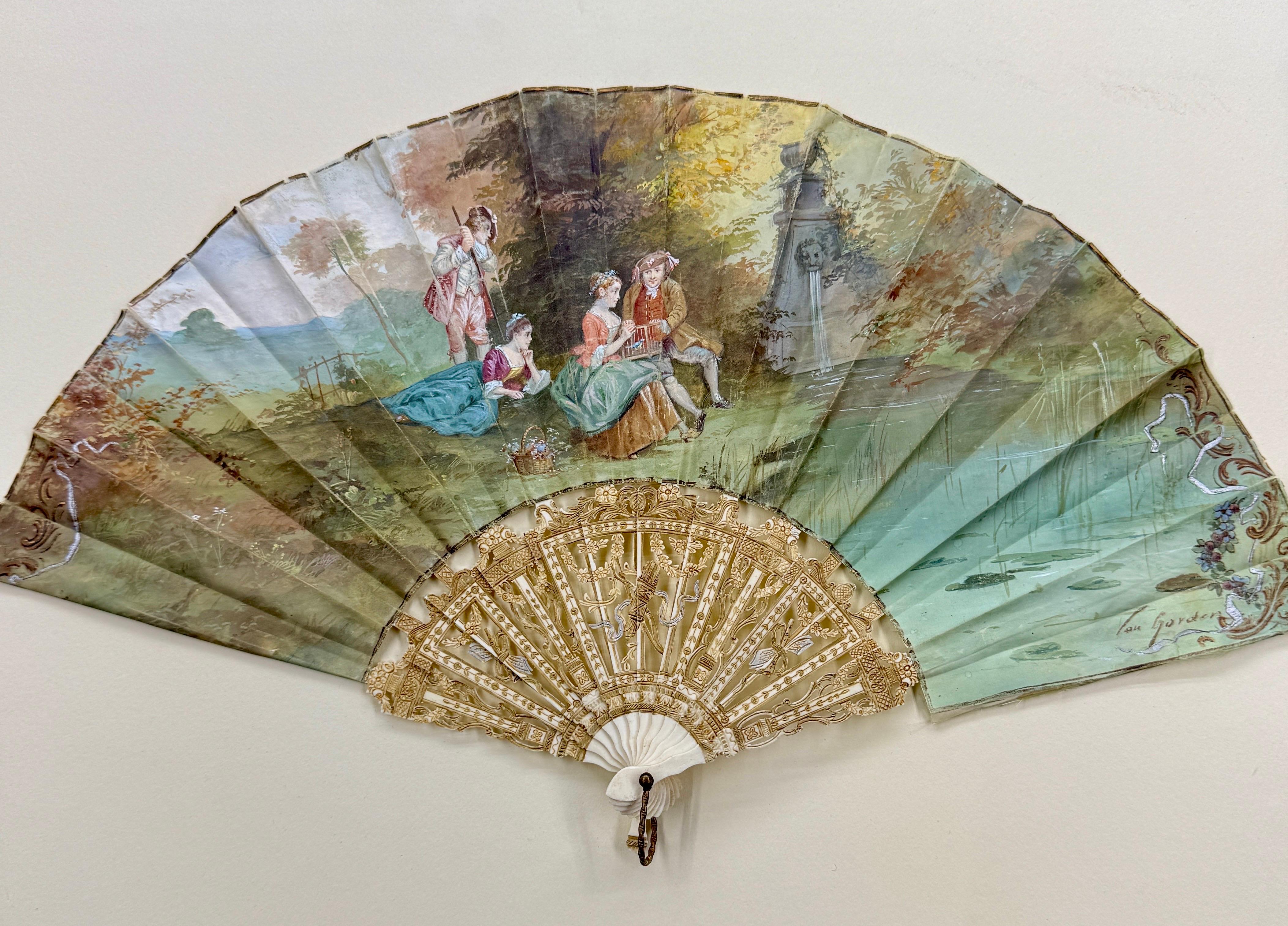 Impressive hand fan with a painted pastoral scene. The scene depicts figures enjoying leisurely activities in a picturesque landscape. The paper appears to be a textured woven silk. The sticks or ribs are carved and pierced. The fan is mounted on