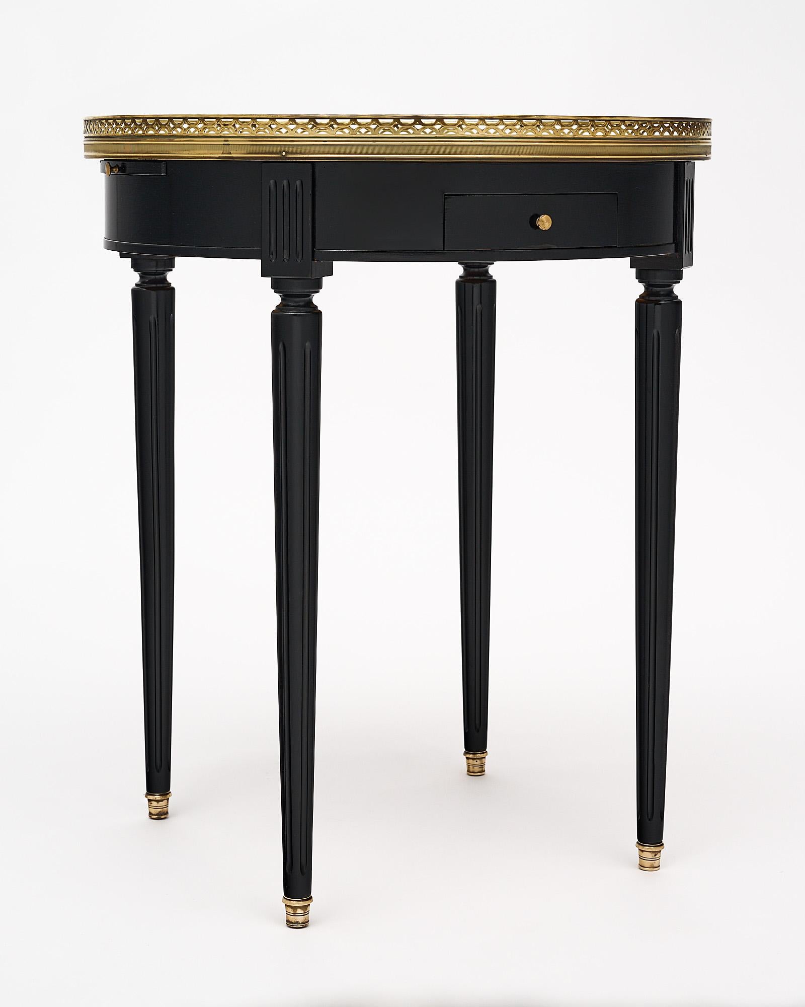 Side table, French, made of mahogany that has been finished in an ebony French polish with a Carrara marble top. The top is framed in an opened brass gallery. There are two small dovetailed drawers and two small leaves with green leather tops.