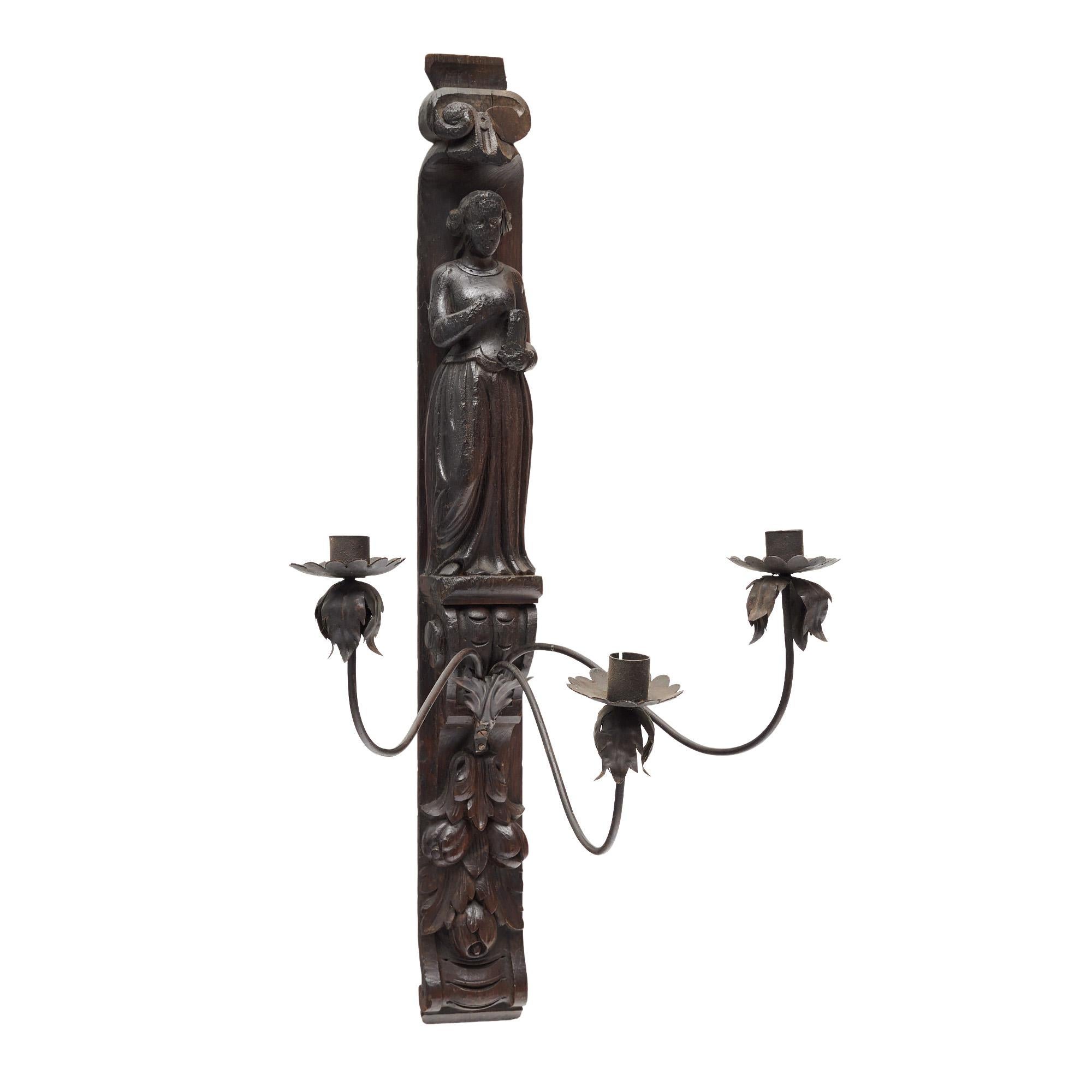 Probabilmente un frammento di un pannello di una chiesa francese, questa applique in legno intagliato a mano proveniente dalla Francia presenta un candelabro in ferro che contiene tre candele. Il legno è intagliato con l'immagine di una donna e un