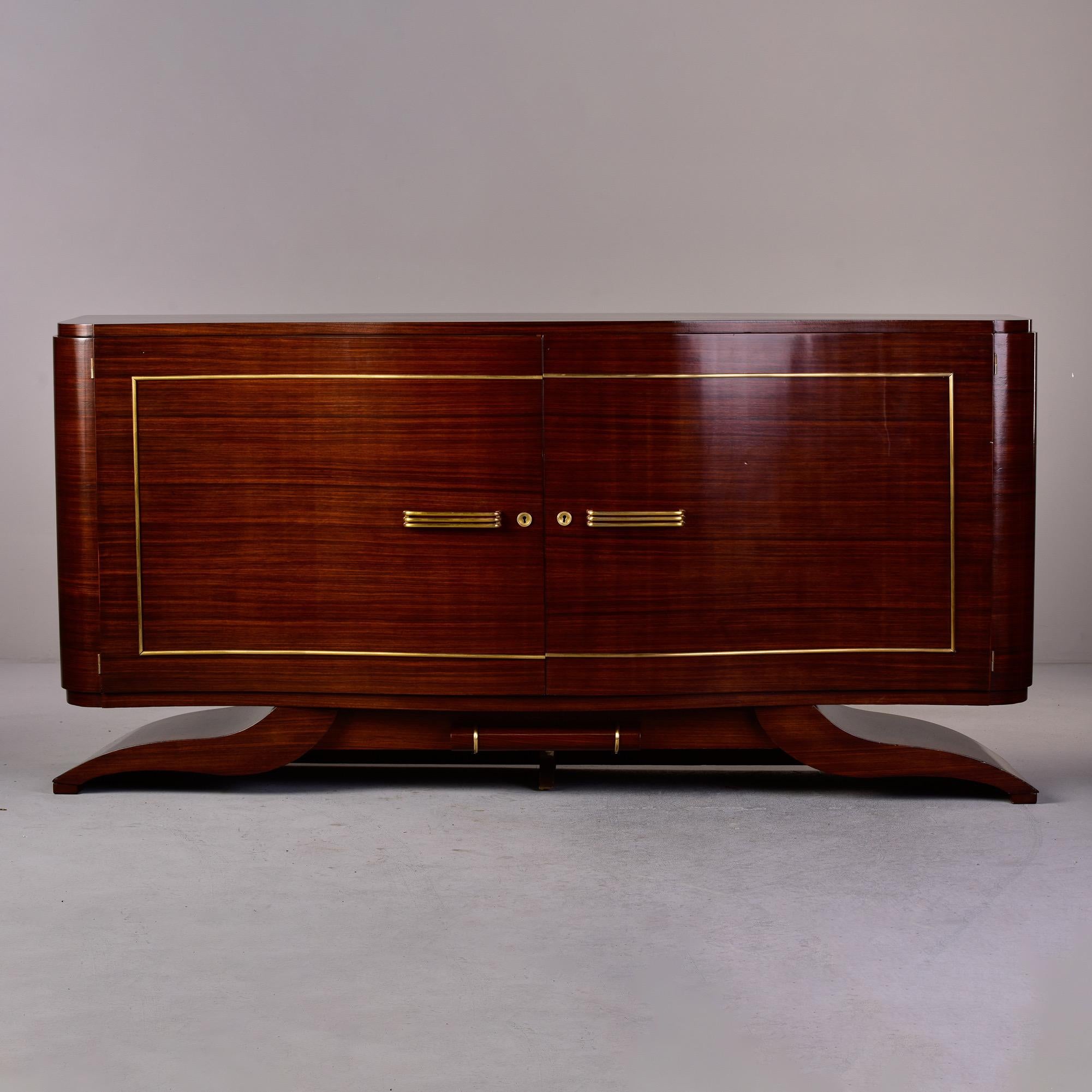Circa 1940s French mahogany buffet with sleek, curvy Art Deco lines and brass trim. Locking cabinet opens to a slim top drawer on each side and storage with a single internal shelf on each side. Unknown maker.