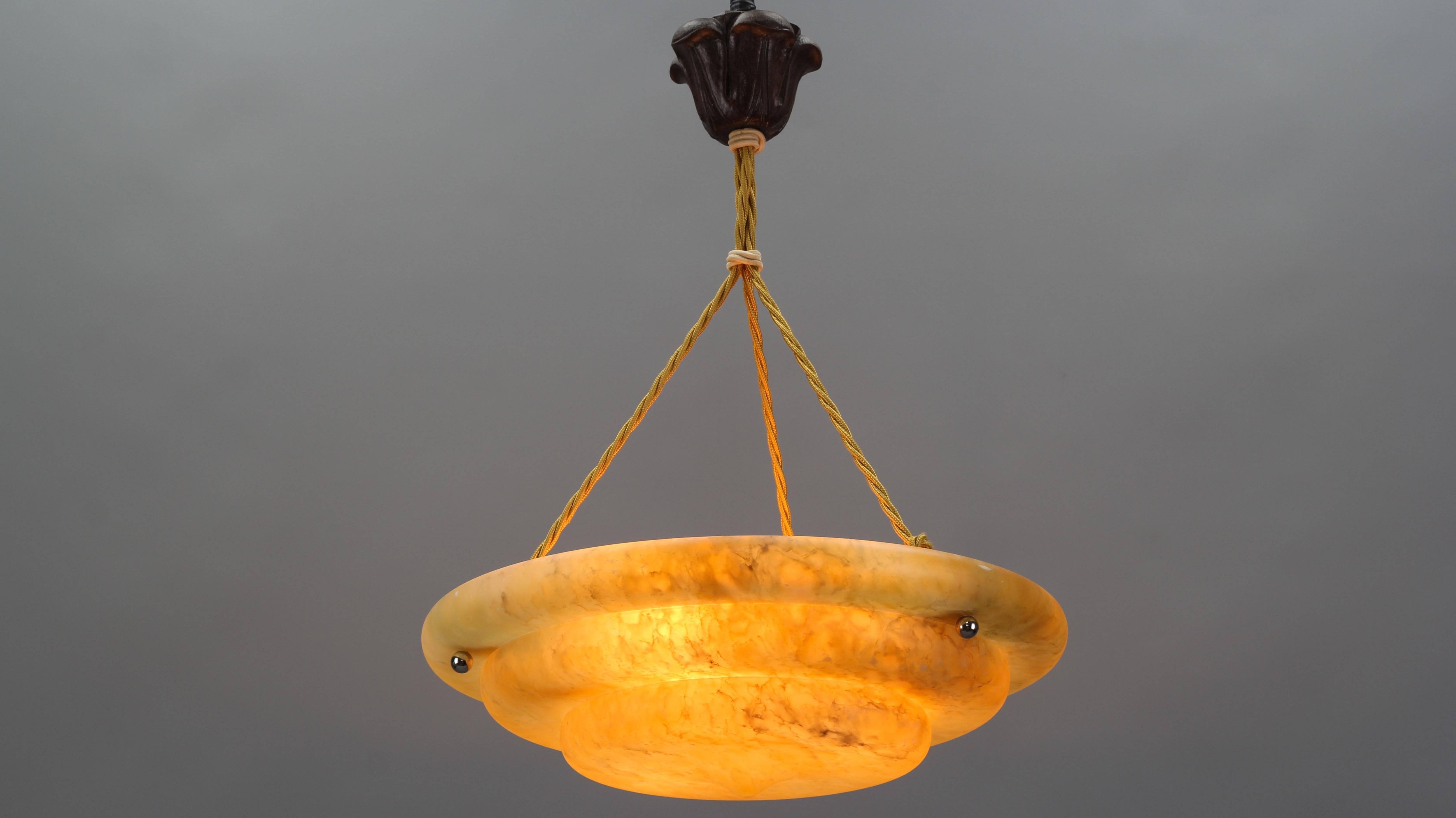 Beautifully shaped French Art Deco style alabaster pendant ceiling light fixture in warm amber color. The masterfully carved one-piece alabaster bowl is suspended by three ropes and a wooden ceiling rose. The light shining through the stone with