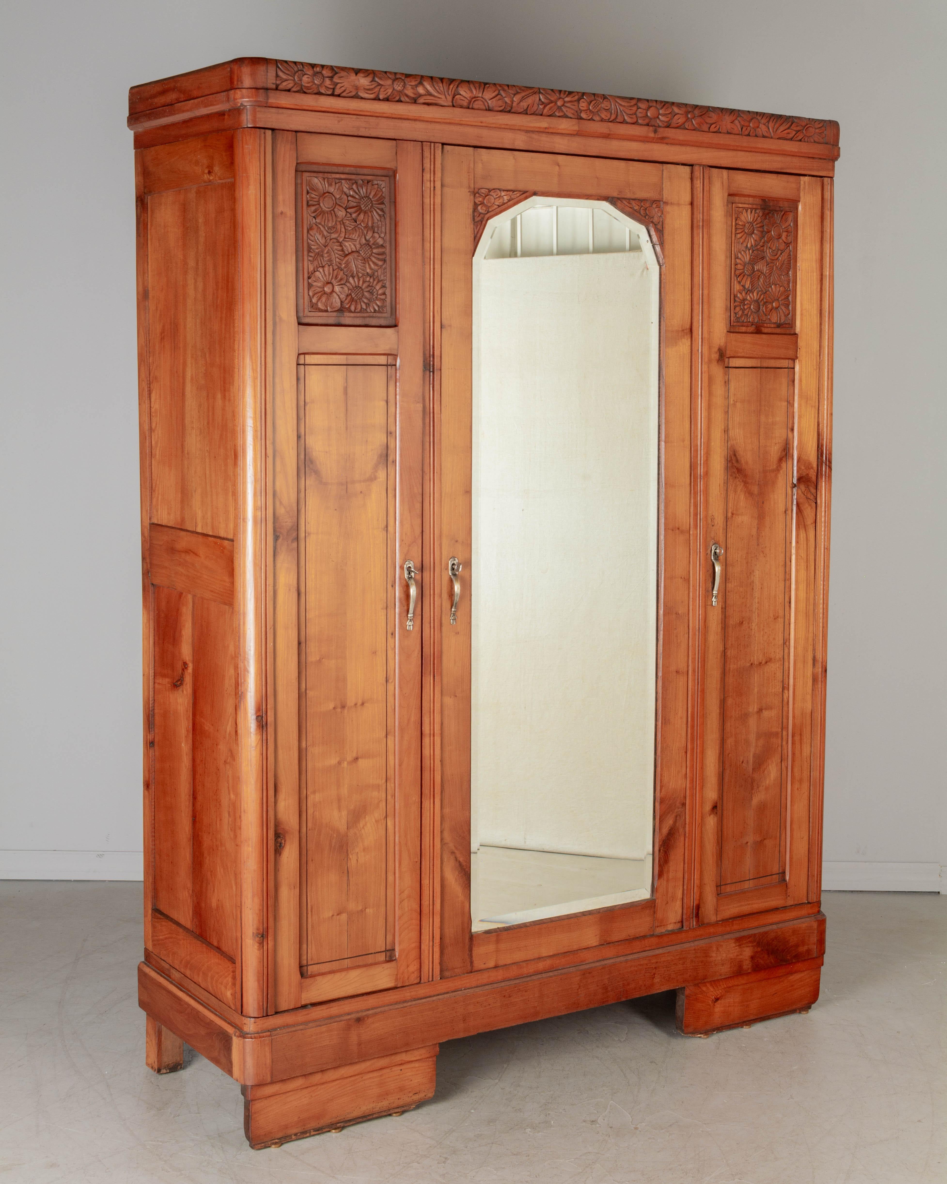 A French Art Deco three-door armoire made of solid cherry wood, nicely detailed with hand-carved stylized floral panels at the top. Center door with original beveled mirror. Nickel plated cast brass hardware with working locks and three keys. Right