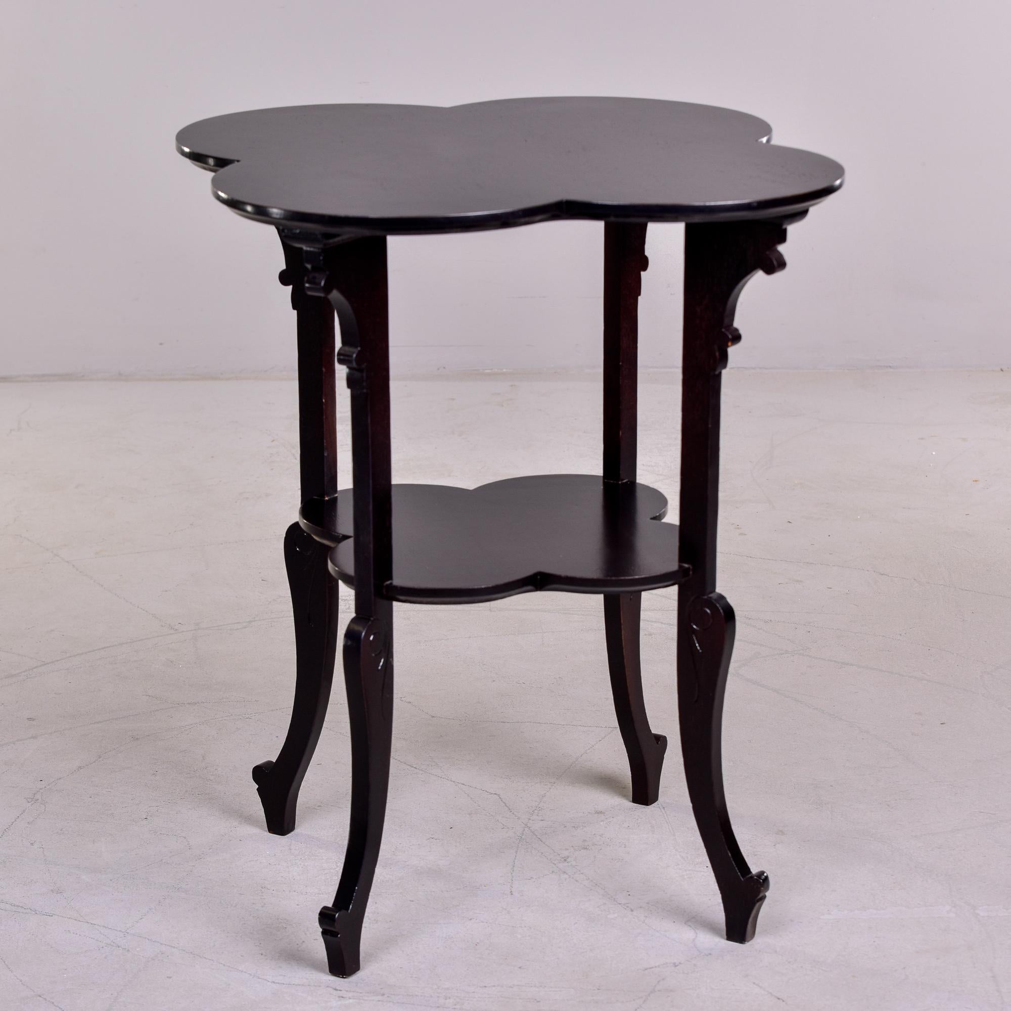 Circa 1930s French side table of black painted oak has a four leaf clover or quatrefoil shaped top and lower shelf and cabriole legs with carved details at the knees and top supports. Unknown maker.