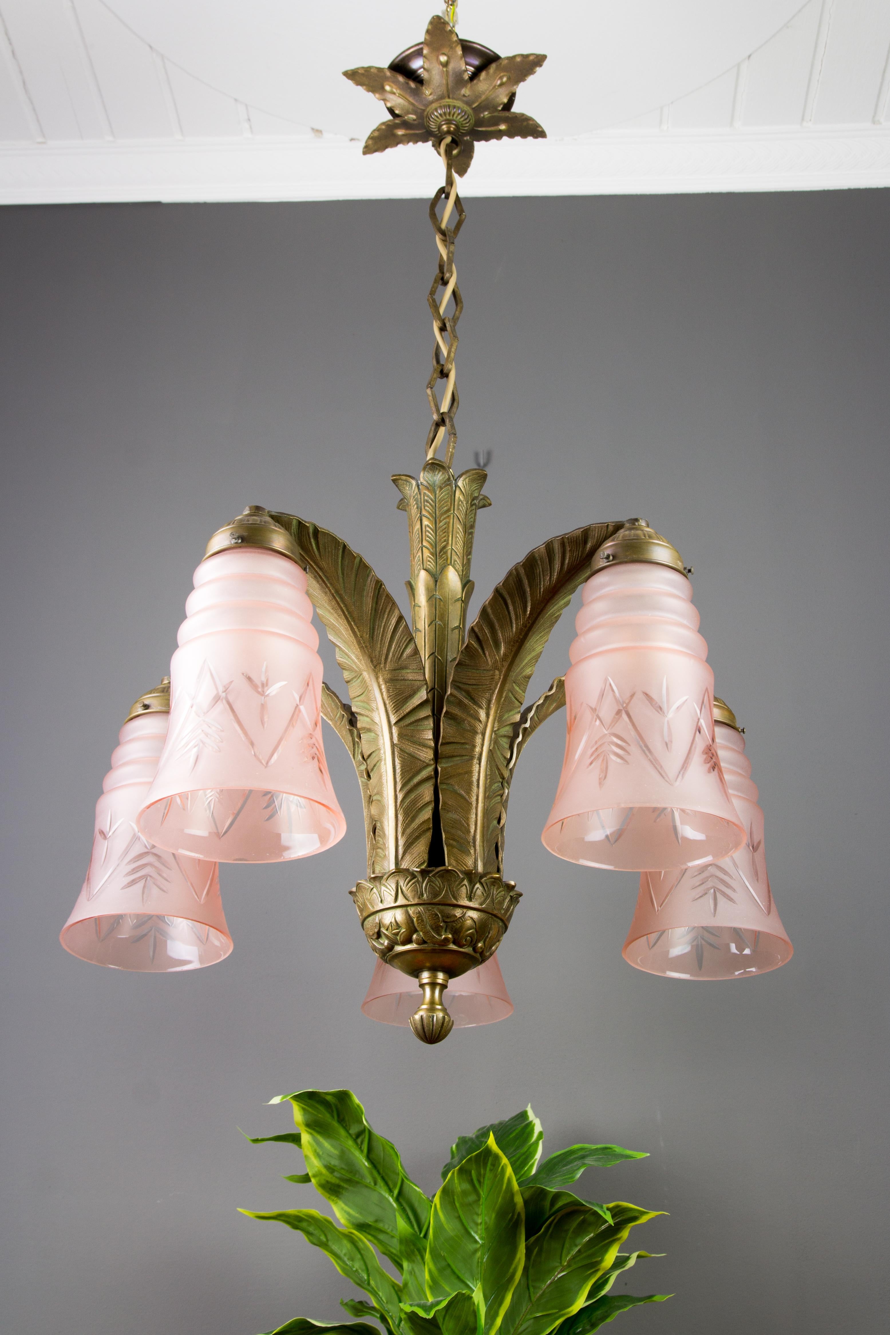 An adorable French Art Deco bronze chandelier, made in the 1930s. Five large bronze arms in the shape of big leaves are holding five pink frosted bell-shaped glass shades with engraved geometrical ornaments. 
The canopy is trimmed with flower