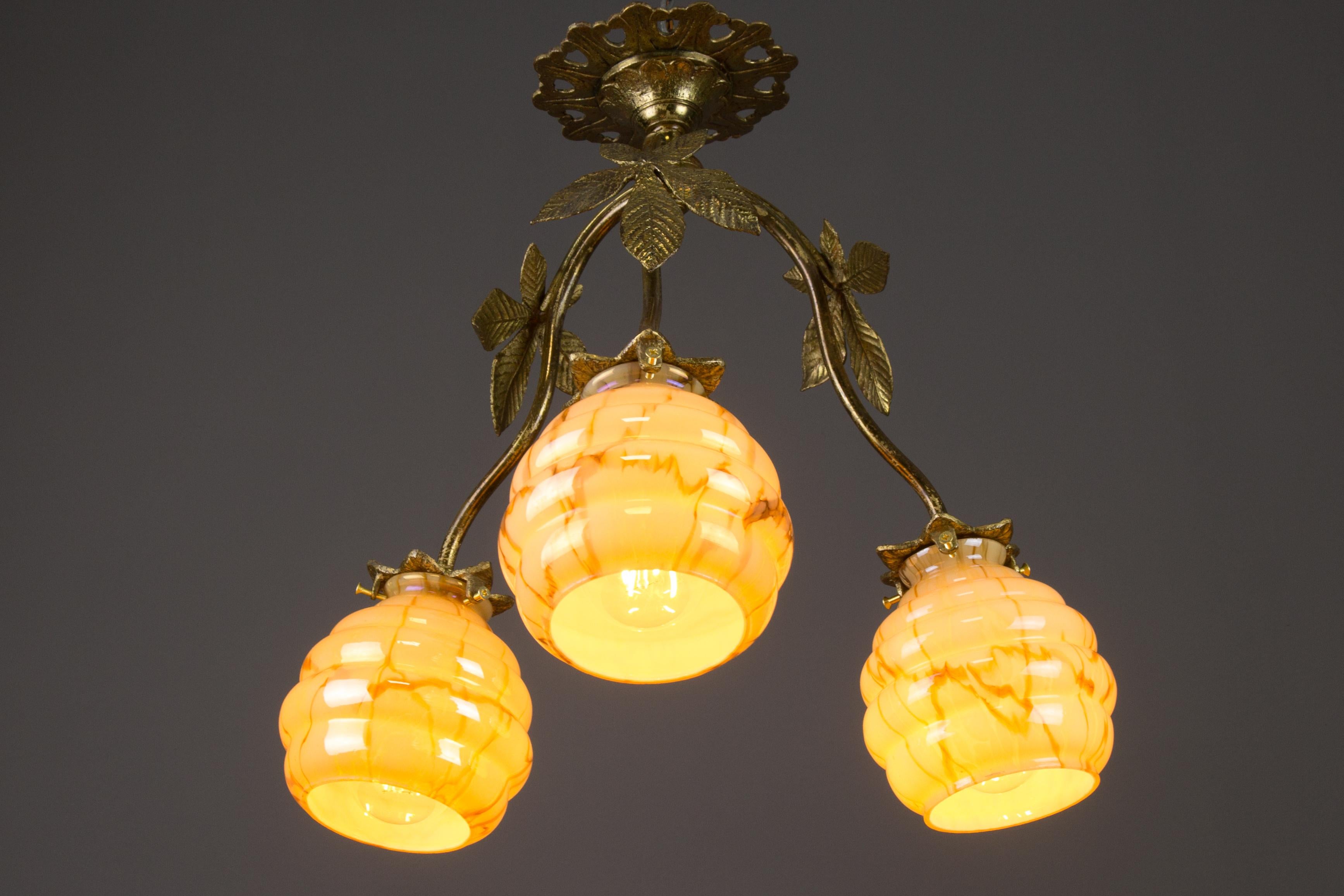 Beautiful French Art Deco bronze and brass chandelier with chestnut leaves and three arms, each with an amber-colored, marbled, and chestnut-shaped glass shade. Adorable beige and golden-colored glass when unlit. When lit, warm and amber-colored