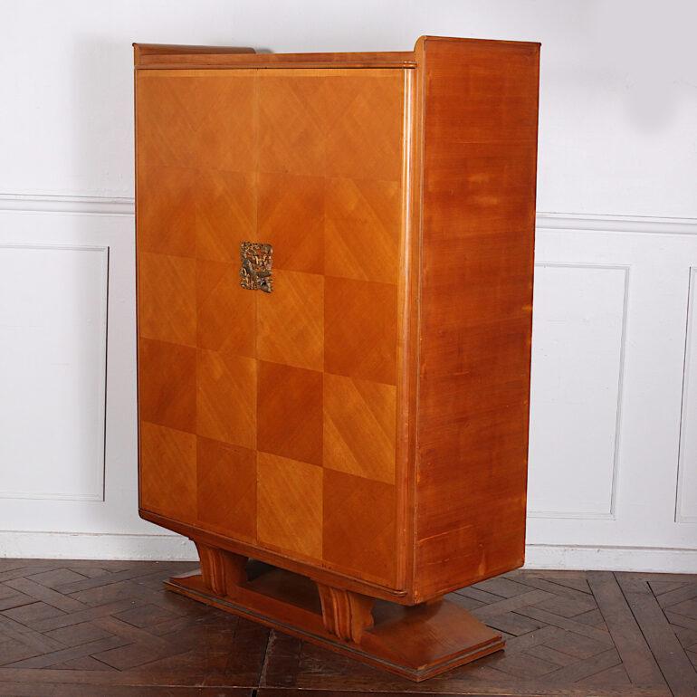 A French Art Deco two door cabinet with cherry-veneered chequer-board parquetry front and gilt bronze figural key plate, the whole raised on simple geometric legs above a plinth base with metal trim. Adjustable shelves to the interior. C. 1930 –