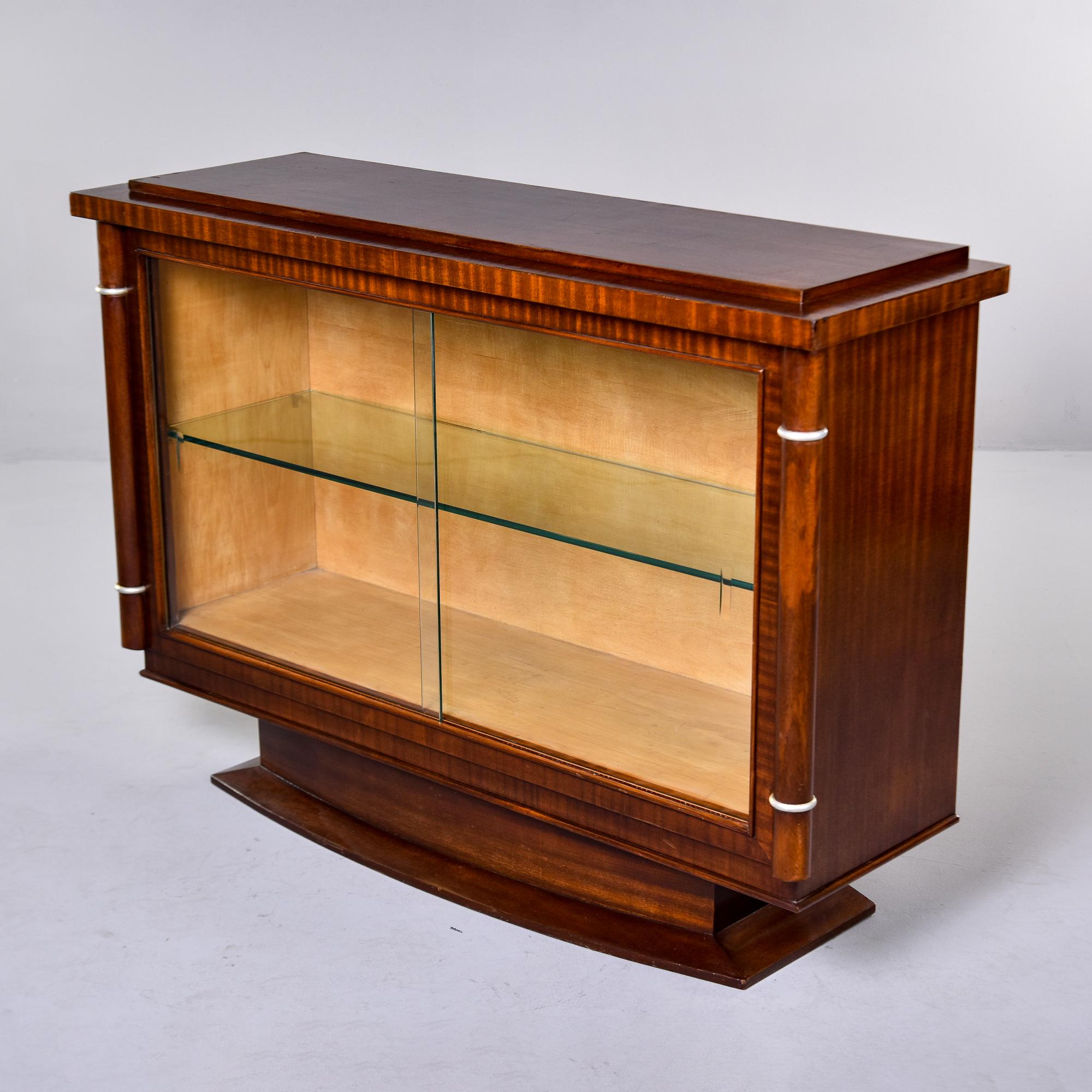 Found in France, this Art Deco cabinet dates from the 1930s. This piece features a wide ovoid pedestal base, sliding glass doors, a single interior fixed glass shelf, decorative columns on the sides and cross banded veneer edge framing the glass