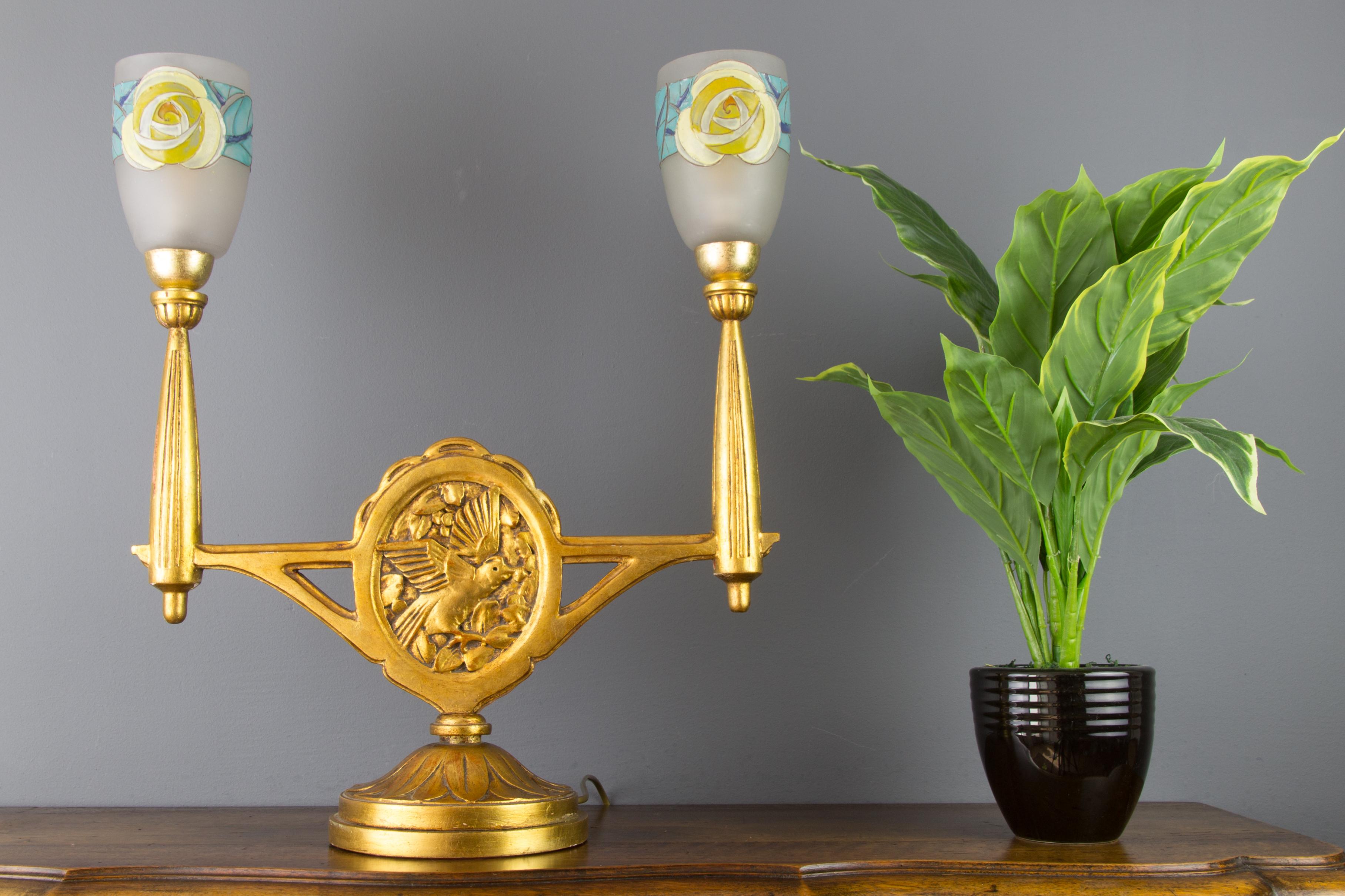 Very unusual and rare Art Deco period table lamp. The base is made of giltwood and has a beautiful carving of birds and leaves. The lamp features two arms, each with an adorable enameled frosted glass shade with floral decors, attributed to Loys